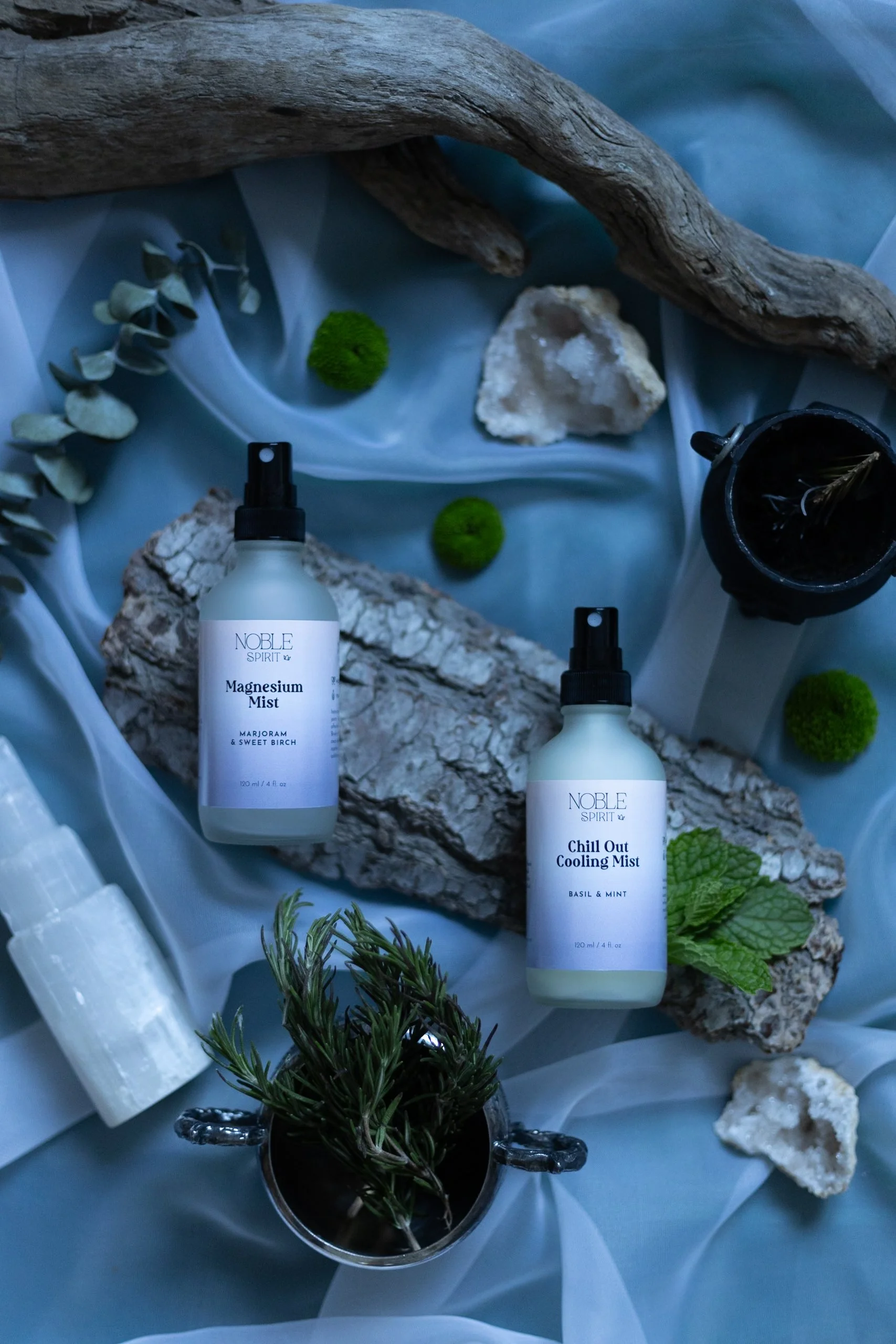 Two spray bottles labeled 'Magnesium Mist' and 'Chill Out Cooling Mist' surrounded by rocks, greenery, driftwood, and white crystals on a light blue fabric background.