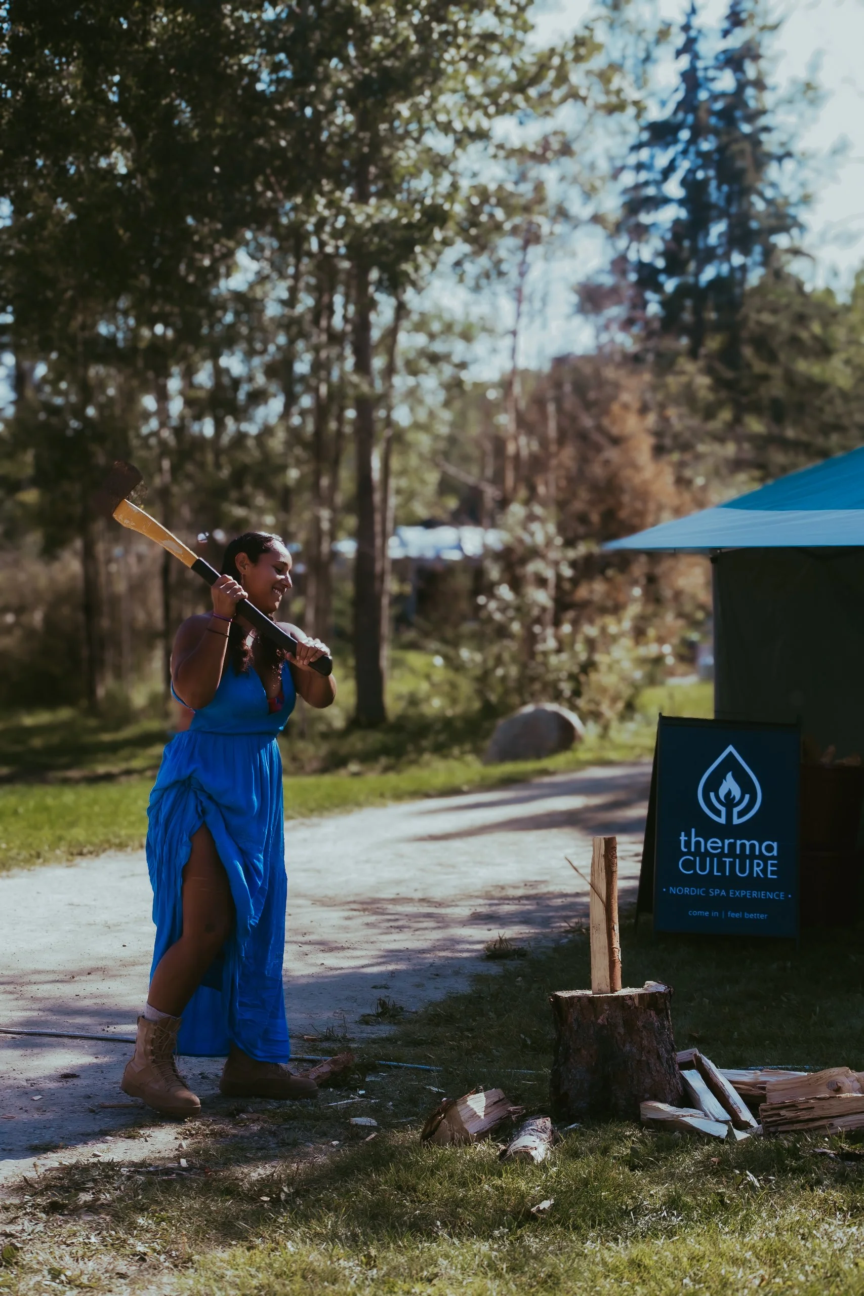 A woman with dark hair in a braid, wearing a blue dress and tan combat boots, holding an axe over her shoulder, standing outdoors near a tent that has a sign reading 'Therma Culture Nordic Spa Experience.' She is smiling and surrounded by trees and l