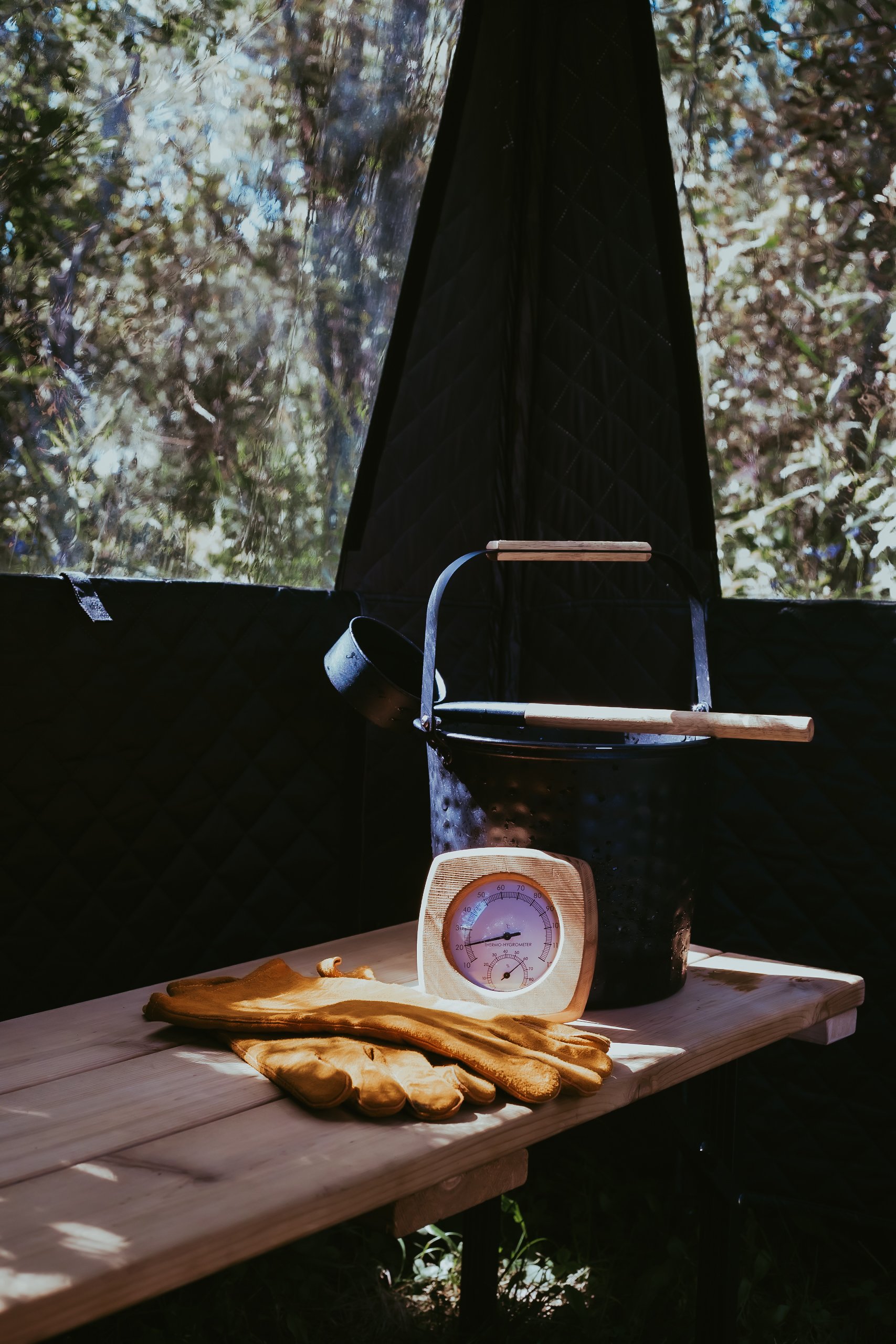 A wooden table with yellow work gloves, a temperature gauge, a black metal bucket, and a long wooden handle tool inside a black insulated tent with transparent window showing trees outside.