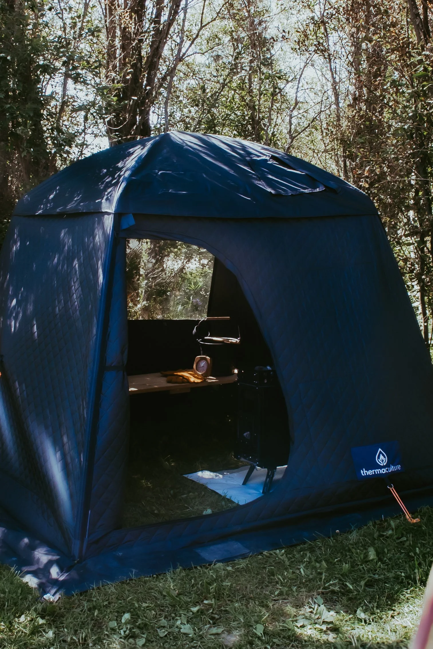 A dark blue thermaculture tent set up in a wooded outdoor area with a small interior table holding a clock and gloves visible through the open door.