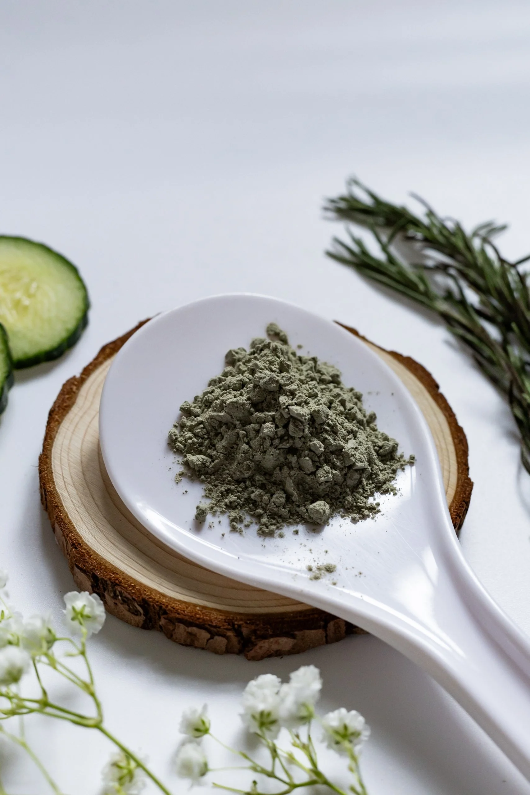 Ground herbal supplement in a white spoon on a wooden block, with cucumber slices, rosemary, and white flowers around.