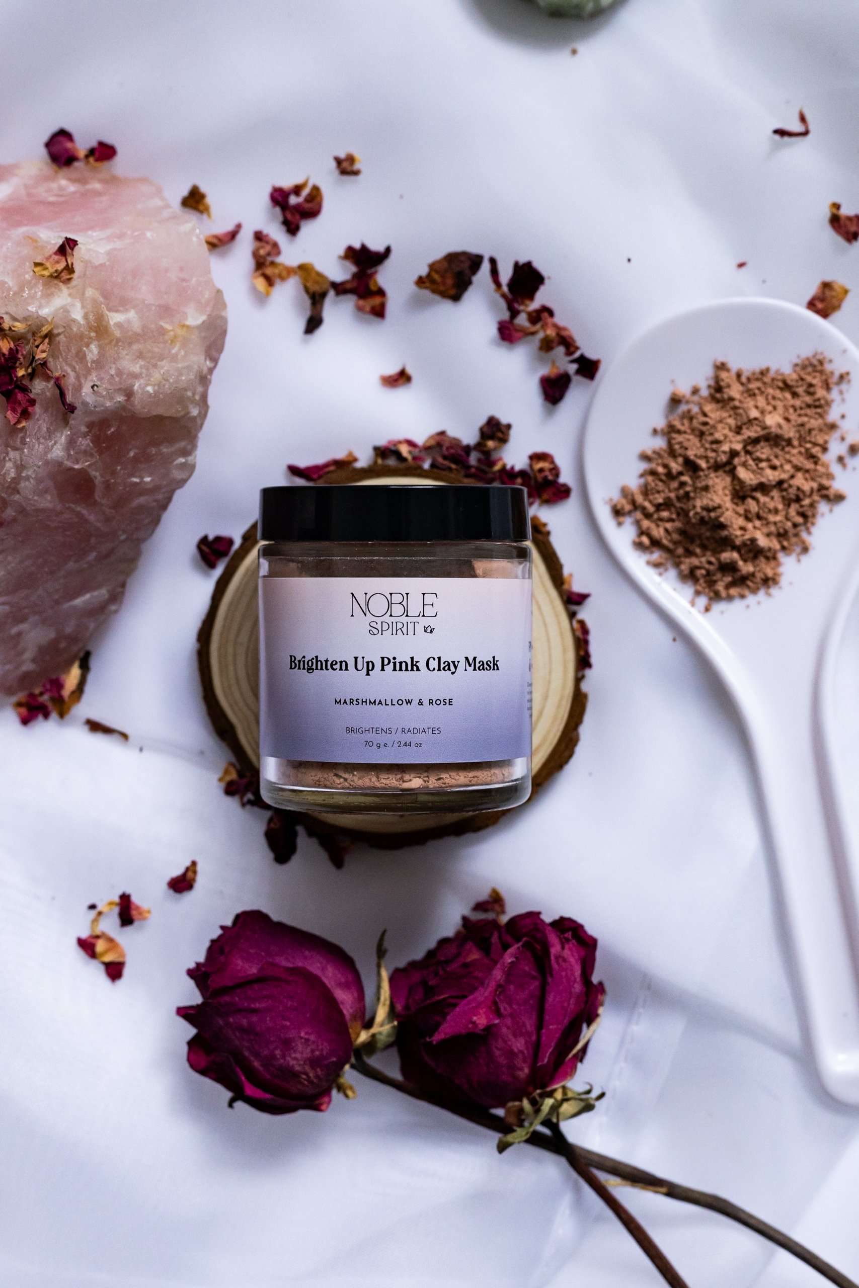 A jar of Brighten Up Pink Clay Mask surrounded by dried roses, rose petals, a large pink salt rock, and small bowls of pink clay powder on a white surface.