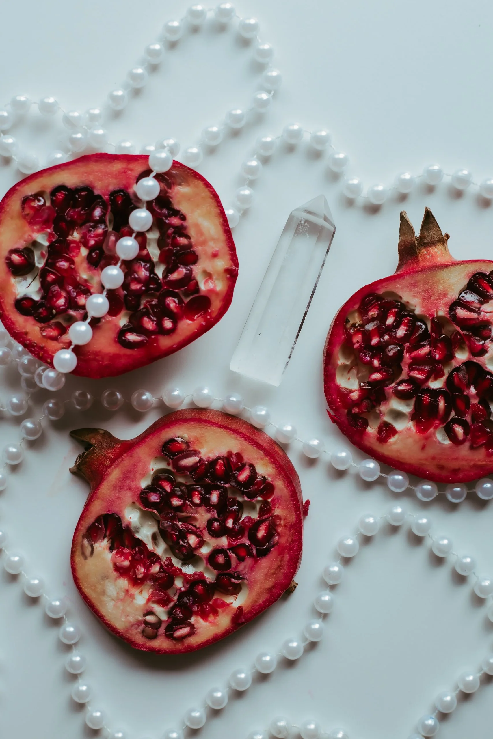 Three halved pomegranates with red seeds, a clear quartz crystal, and pearl necklaces arranged on a white surface.