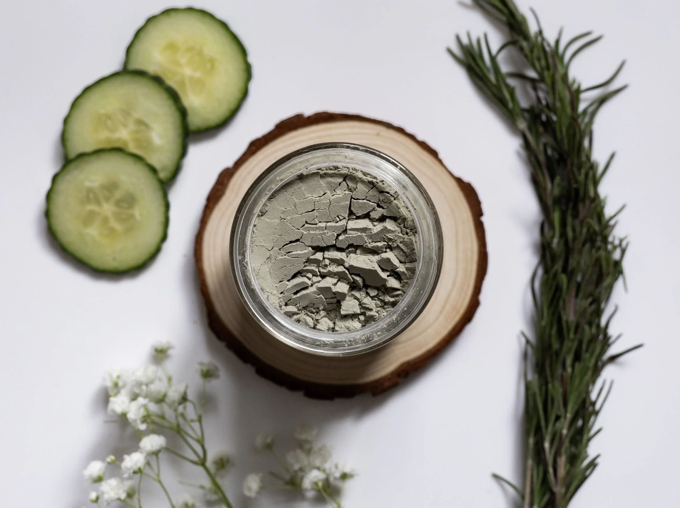 A jar of gray powder on a wooden slab, next to sliced cucumbers, sprigs of rosemary, and white flowers on a white background.