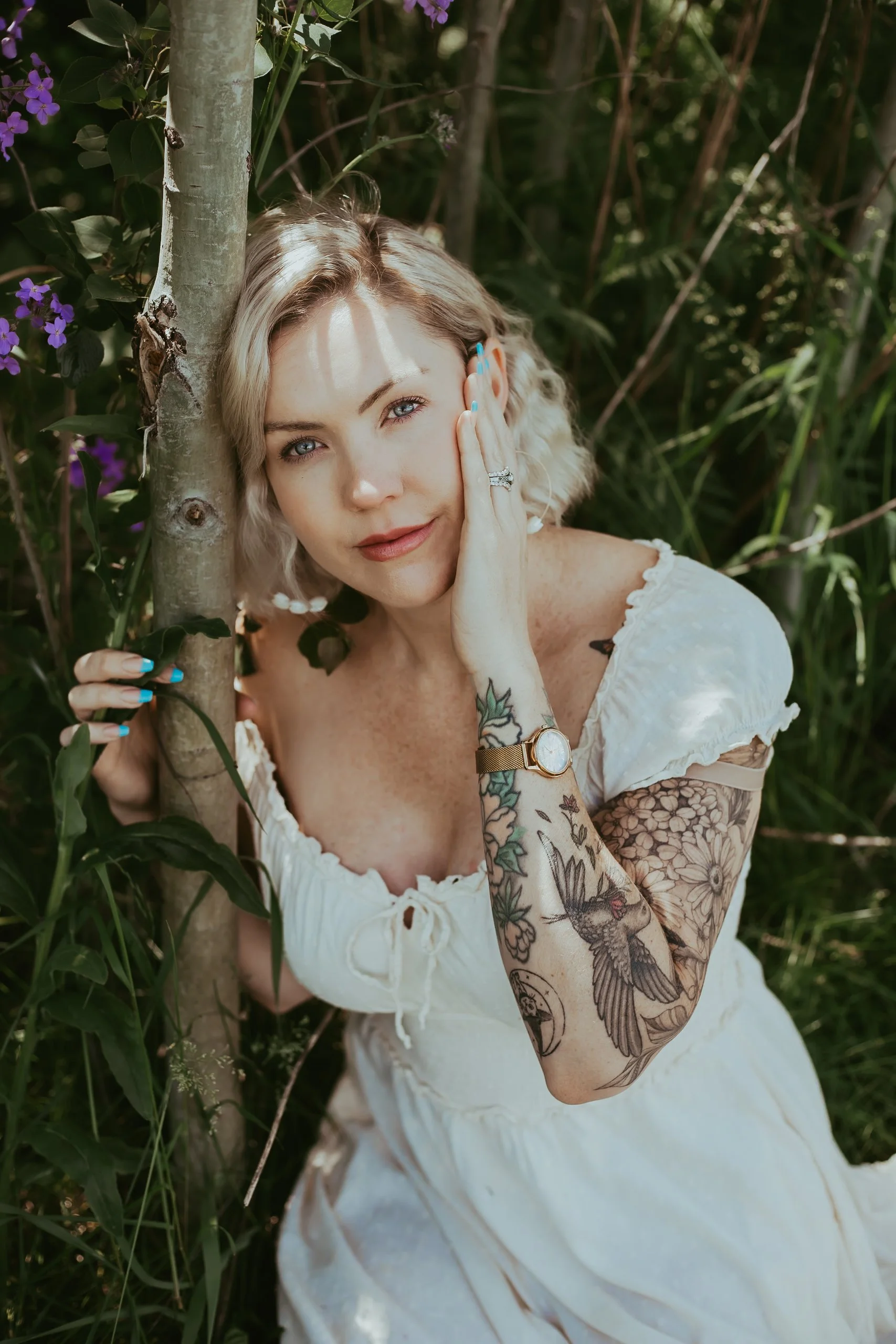 A woman with blonde hair and tattoos on her arms is sitting outdoors among green foliage, holding a tree trunk, and looking at the camera with her hand gently resting on her face.
