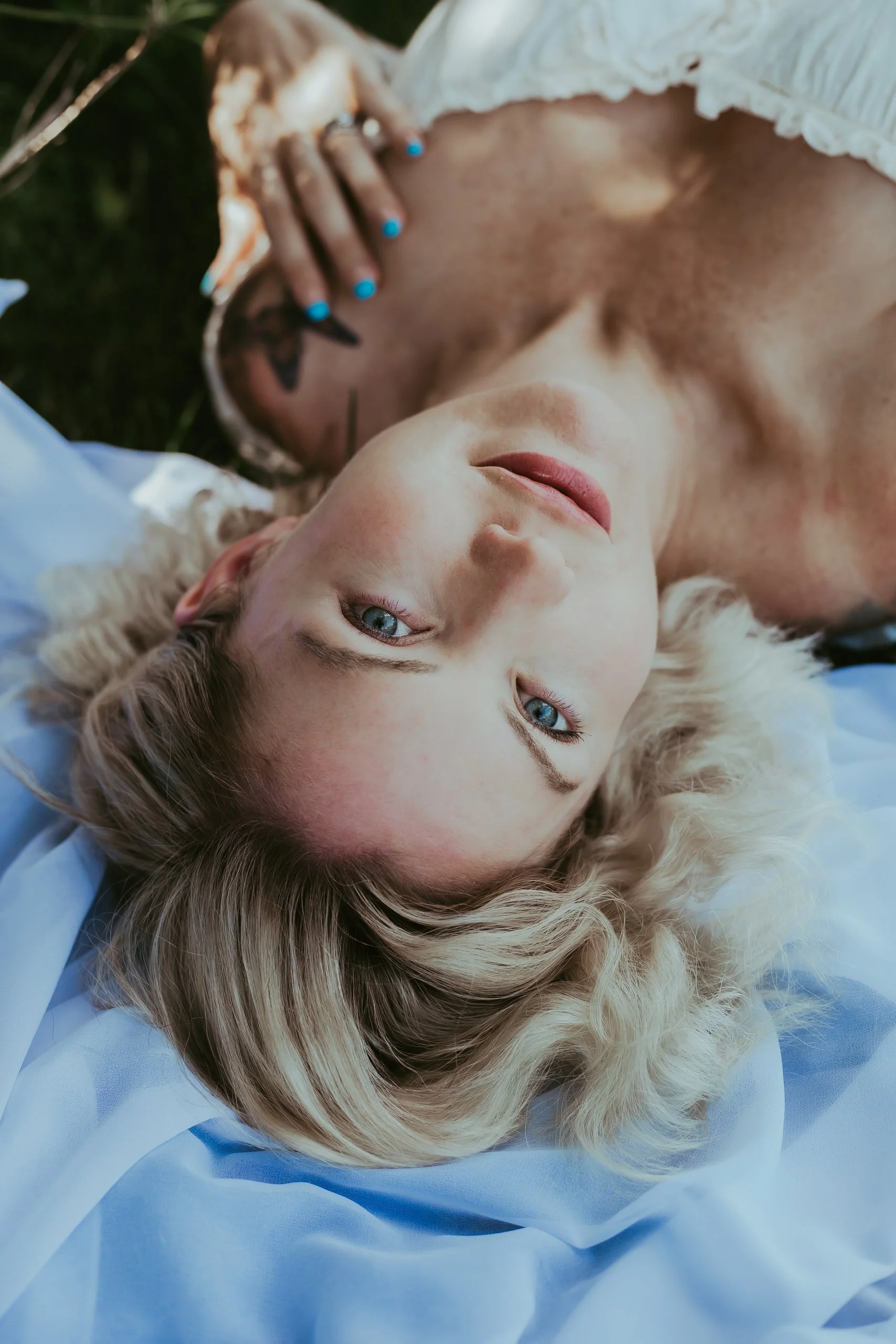 A blonde woman with blue eyes lying on grass with a blue fabric beneath her, looking at the camera, while another person with tattoos covering their face is beside her, partially visible.