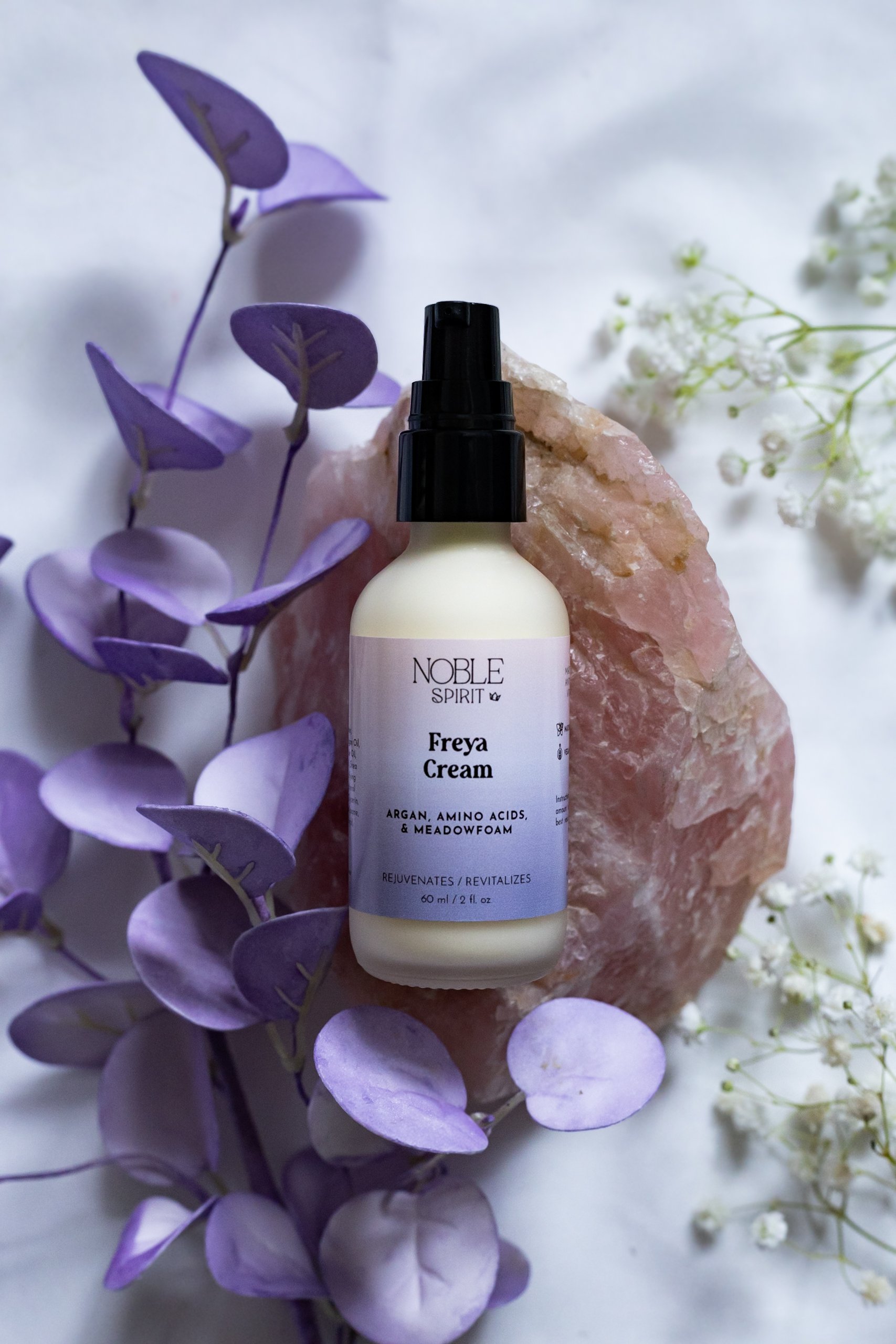 A bottle of Noble Spirit Freya Cream with argan, amino acids, and meadowfoam on a pink rock surrounded by lavender and white flowers.