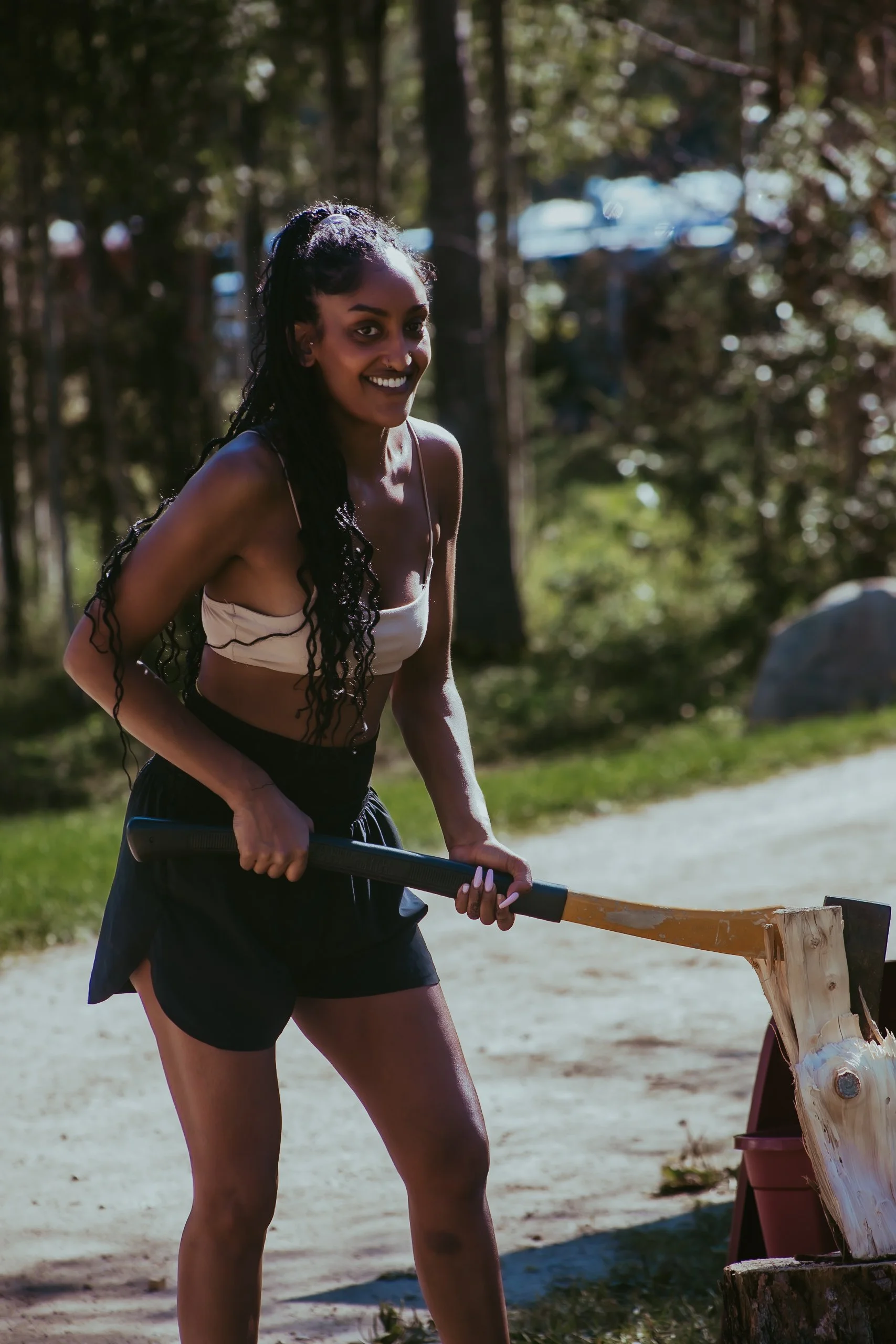 A young woman chopping wood outdoors in a forested area with sunlight filtering through the trees.