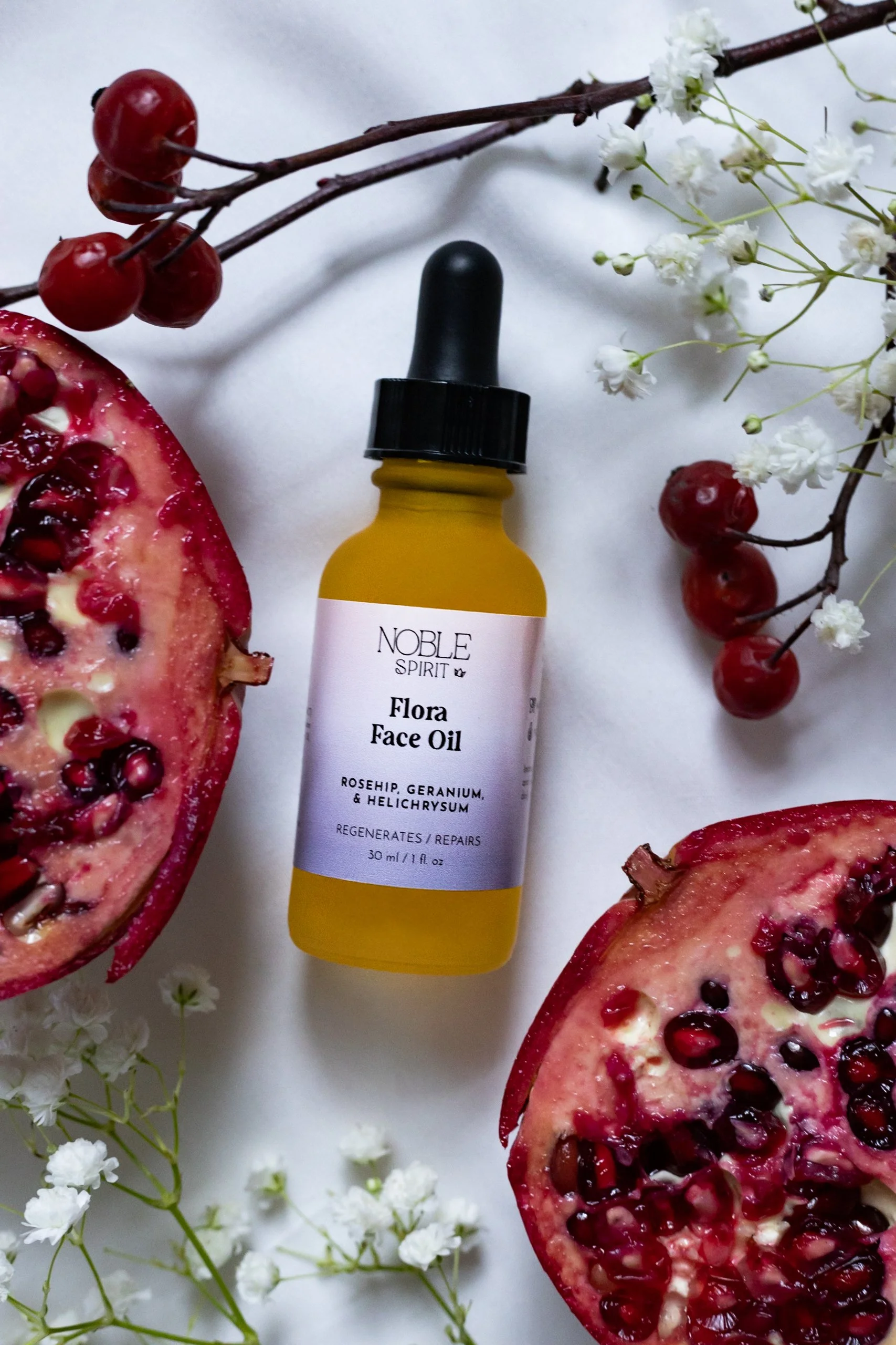 A bottle of Flora Face Oil with rosehip, geranium, and helichrysum, surrounded by pomegranate halves, red berries, and white floral accents on a white background.