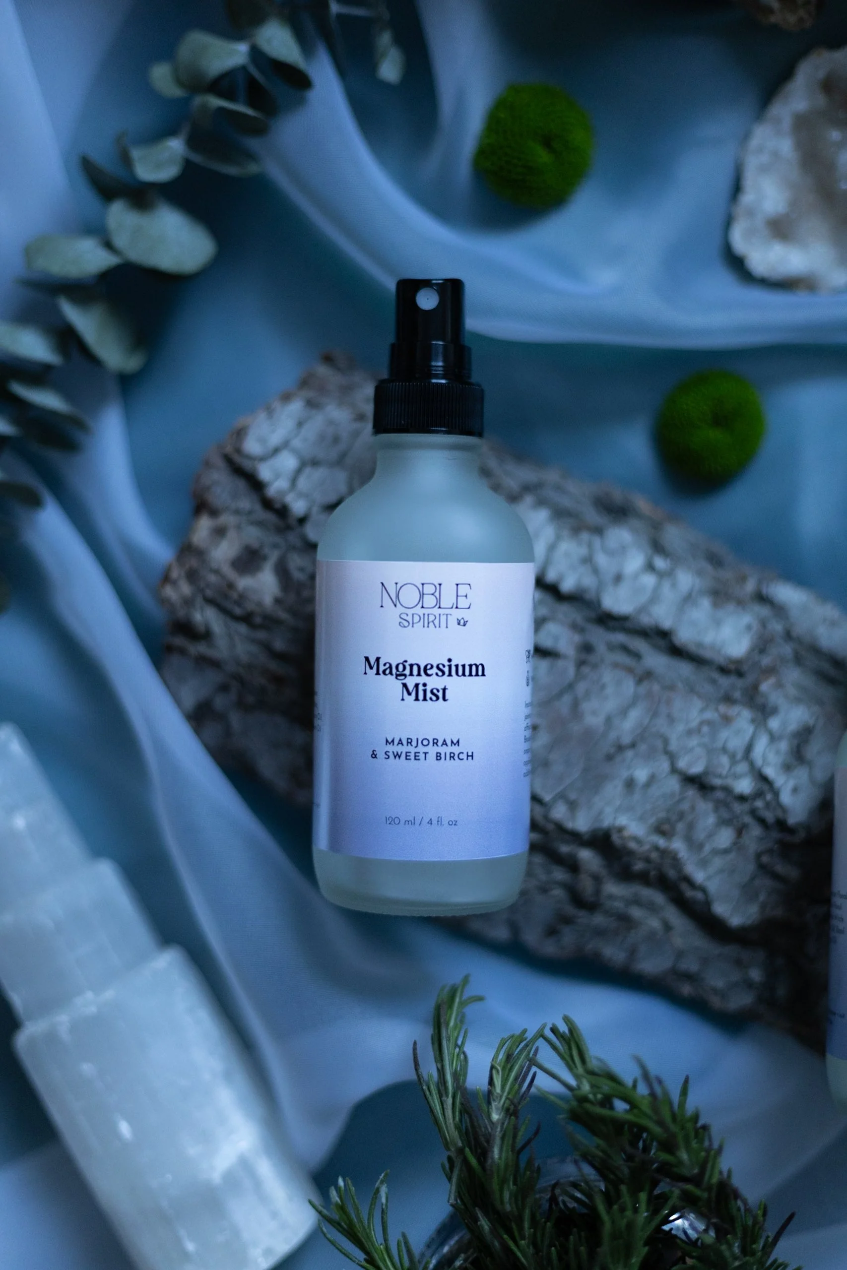 A bottle of Noble Spirit Magnesium Mist with Marjoram and Sweet Birch surrounded by natural elements like moss, moss balls, branches, and rocks.