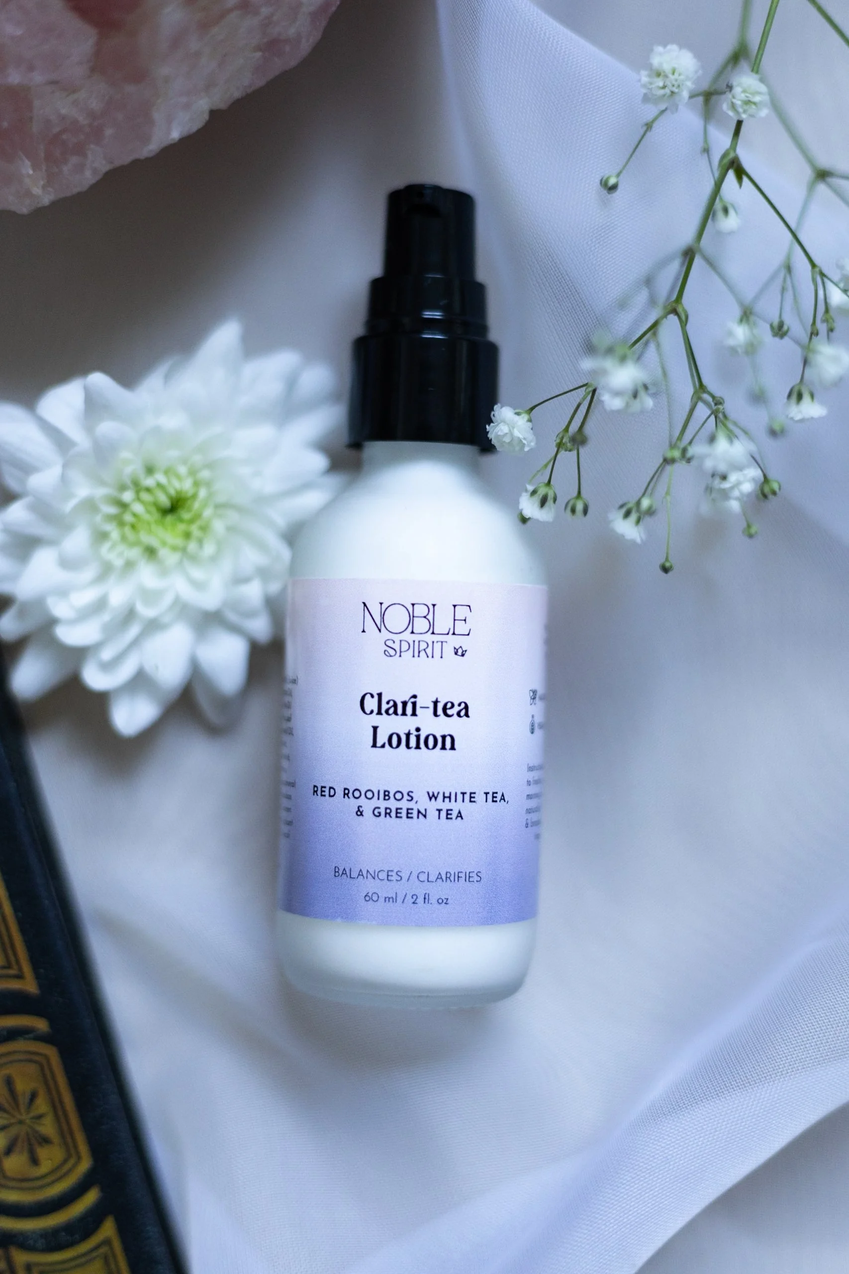 A white spray bottle labeled 'Noble Spirit Clari-Tea Lotion' surrounded by white flowers, including a large dahlia and delicate baby's breath, on a white background.