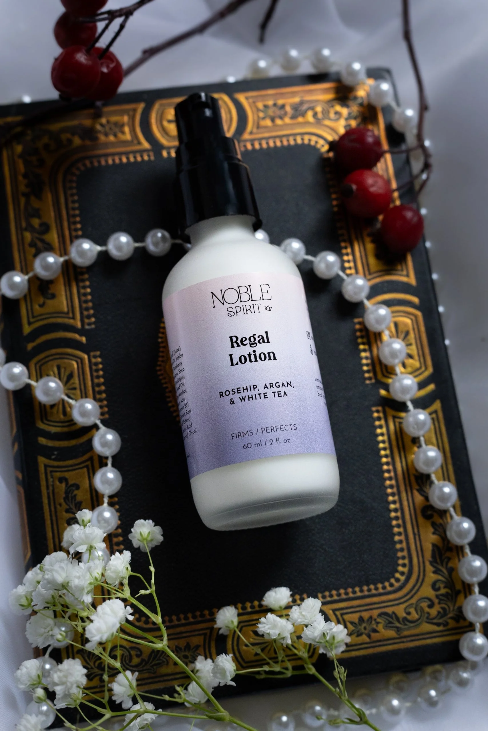 A bottle of Noble Spirit Regal Lotion with rosehip, argan, and white tea on a decorative black and gold tray, surrounded by a string of large white pearls, white baby's breath flowers, and red berries.