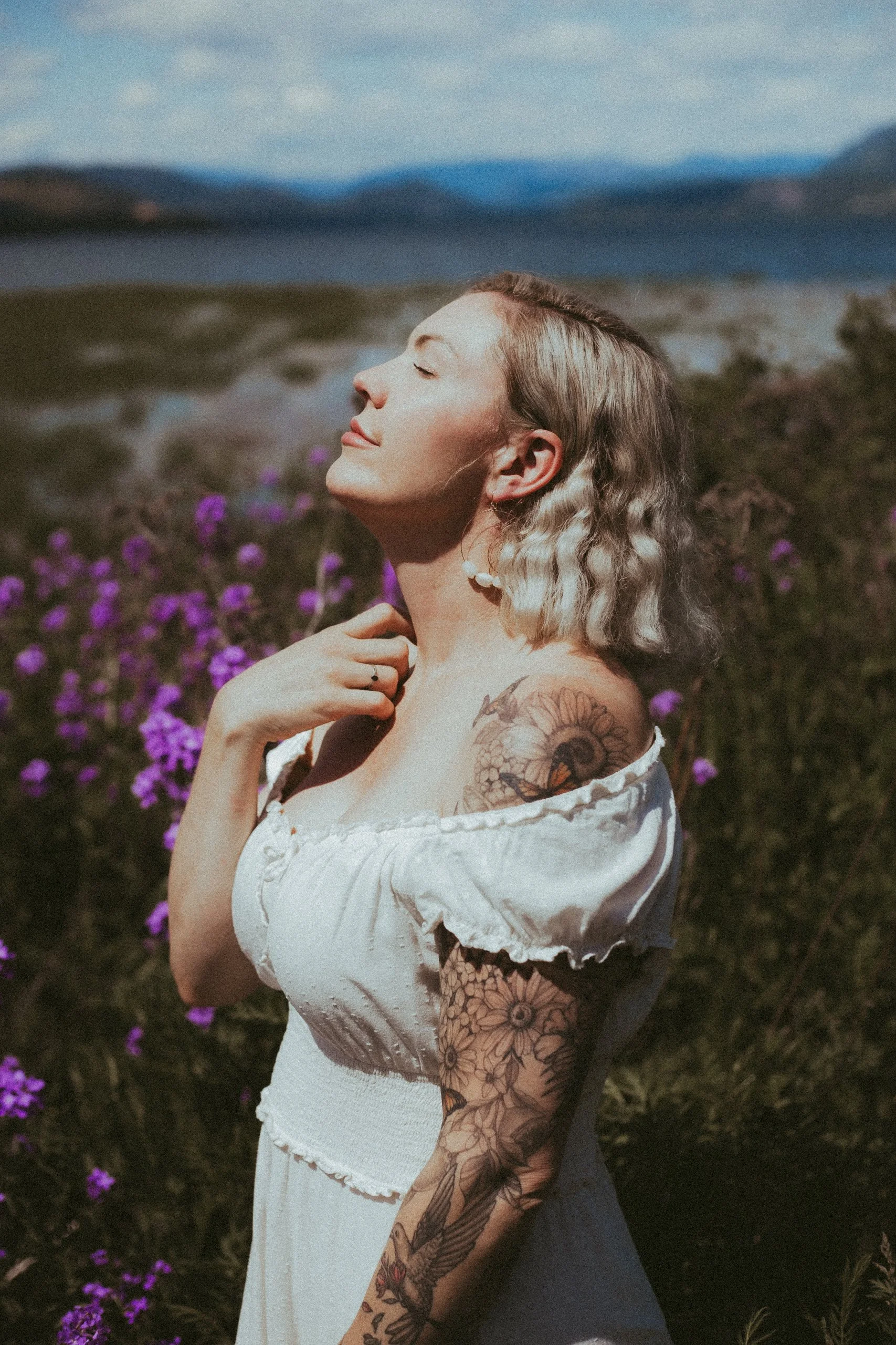 A woman with tattoos and blonde hair standing in a field of purple flowers with her eyes closed and face turned upward, enjoying the sunlight.