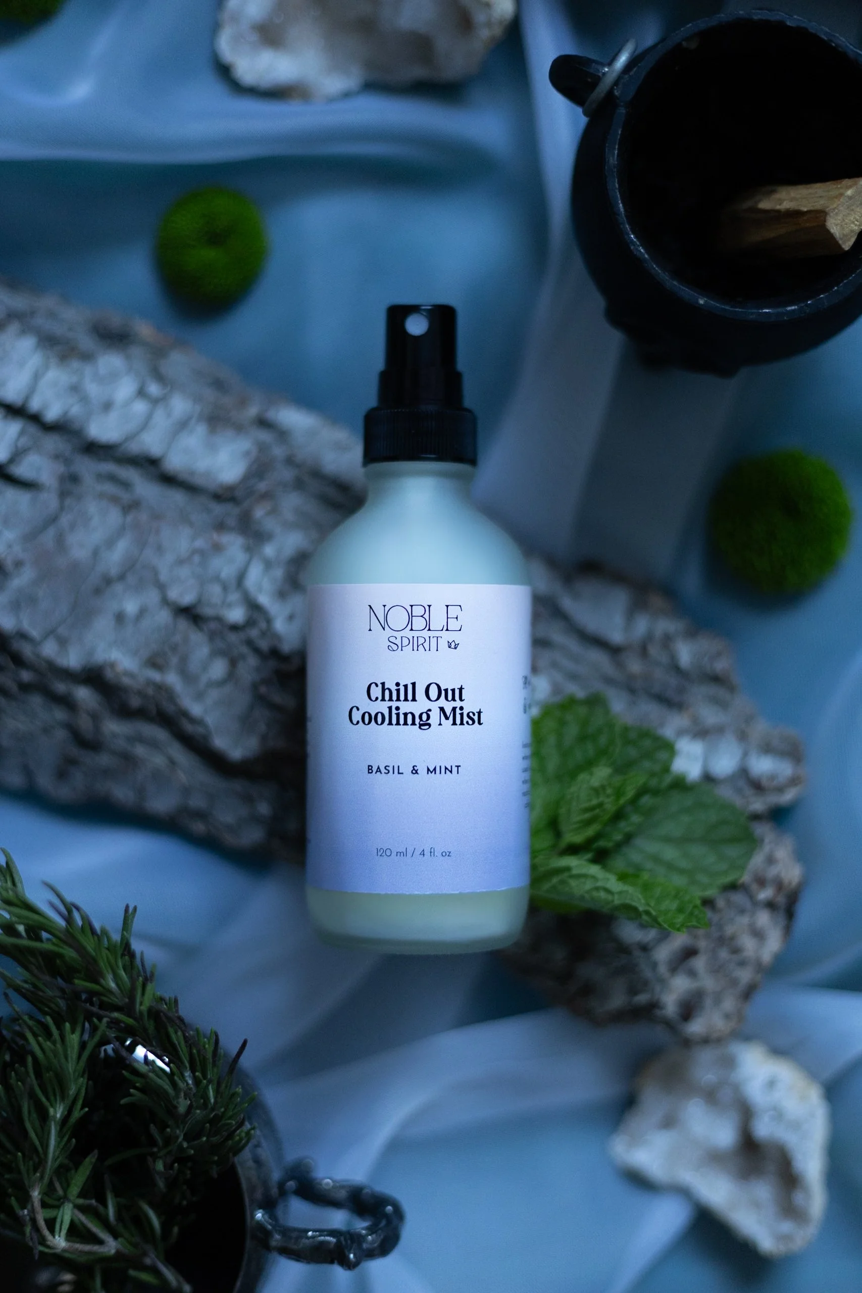 A spray bottle labeled 'Noble Spirit Chill Out Cooling Mist' with basil and mint scent, surrounded by rocks, fresh mint leaves, a piece of driftwood, a small plant, and a black container with wooden sticks, all on a light blue fabric background.