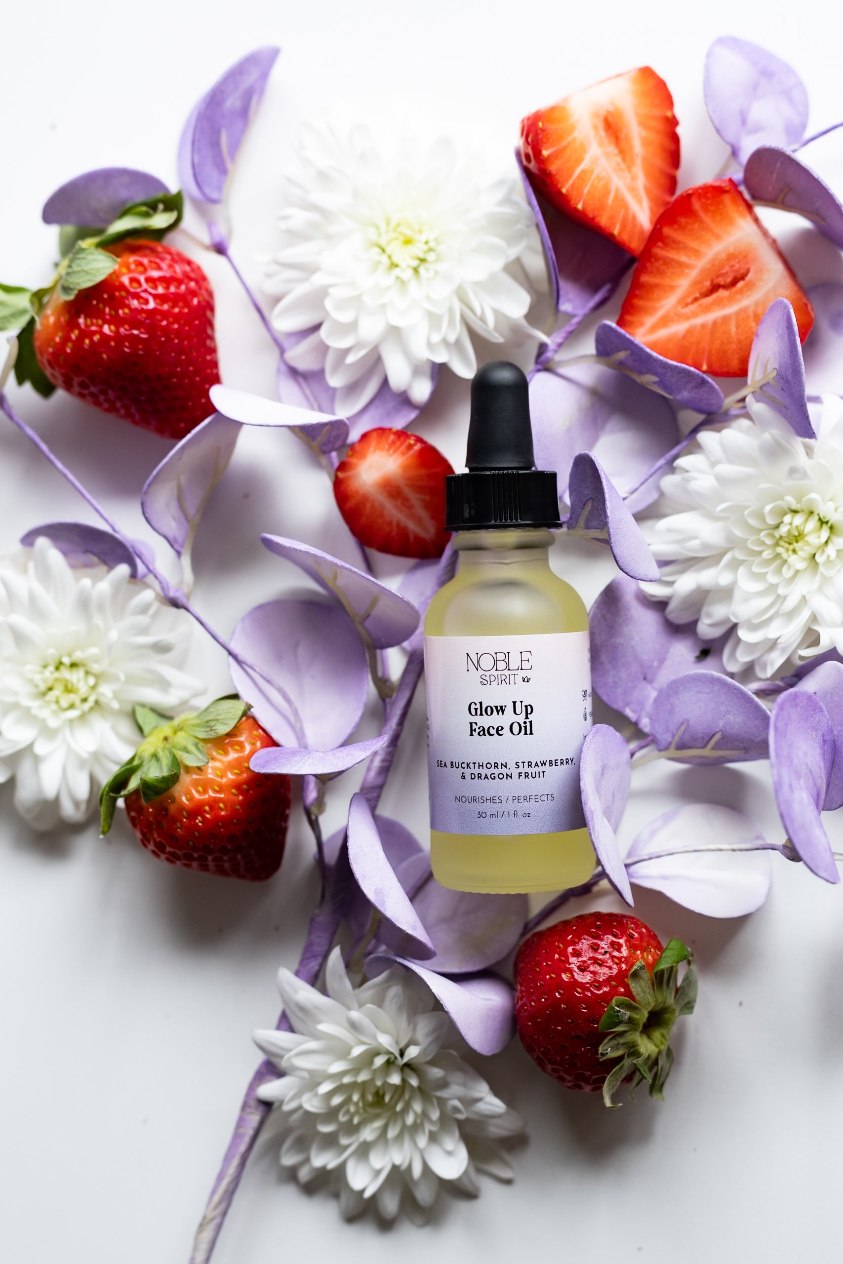 A glass bottle of Noble Spirit Glow Up Face Oil surrounded by strawberries, white chrysanthemums, and purple butterfly-shaped petals on a white background.