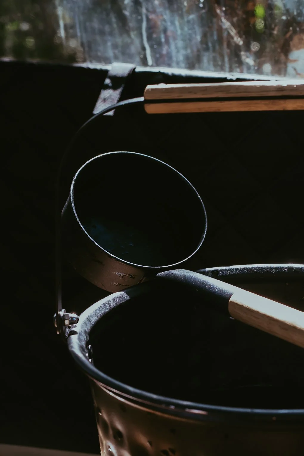 A black bucket and a bucket with a wooden handle are on a dark surface near a window. The window shows a blurry background with trees and sunlight.