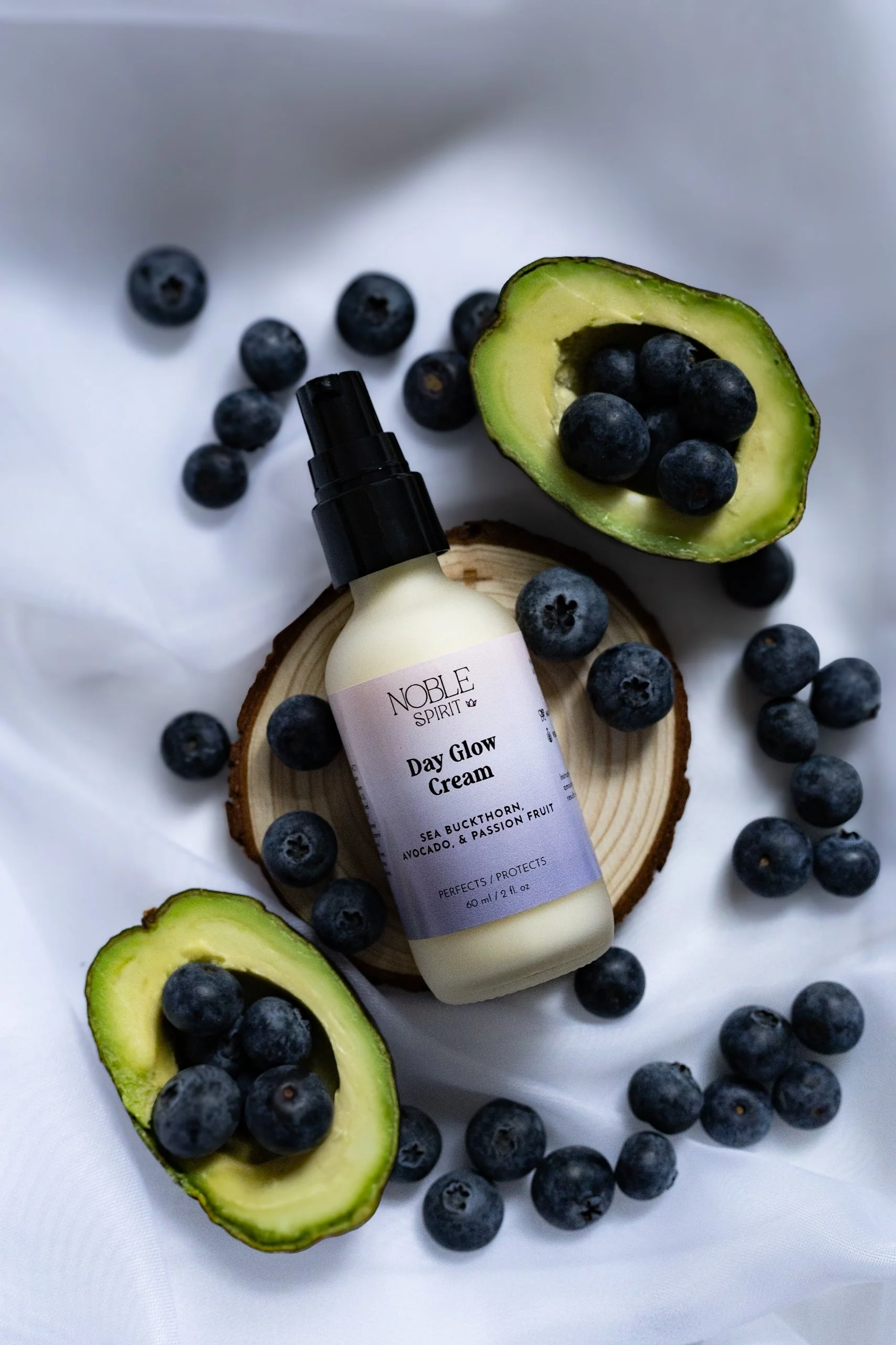 A bottle of Noble Spirit Day Glow Cream surrounded by blueberries and half an avocado on a white surface.