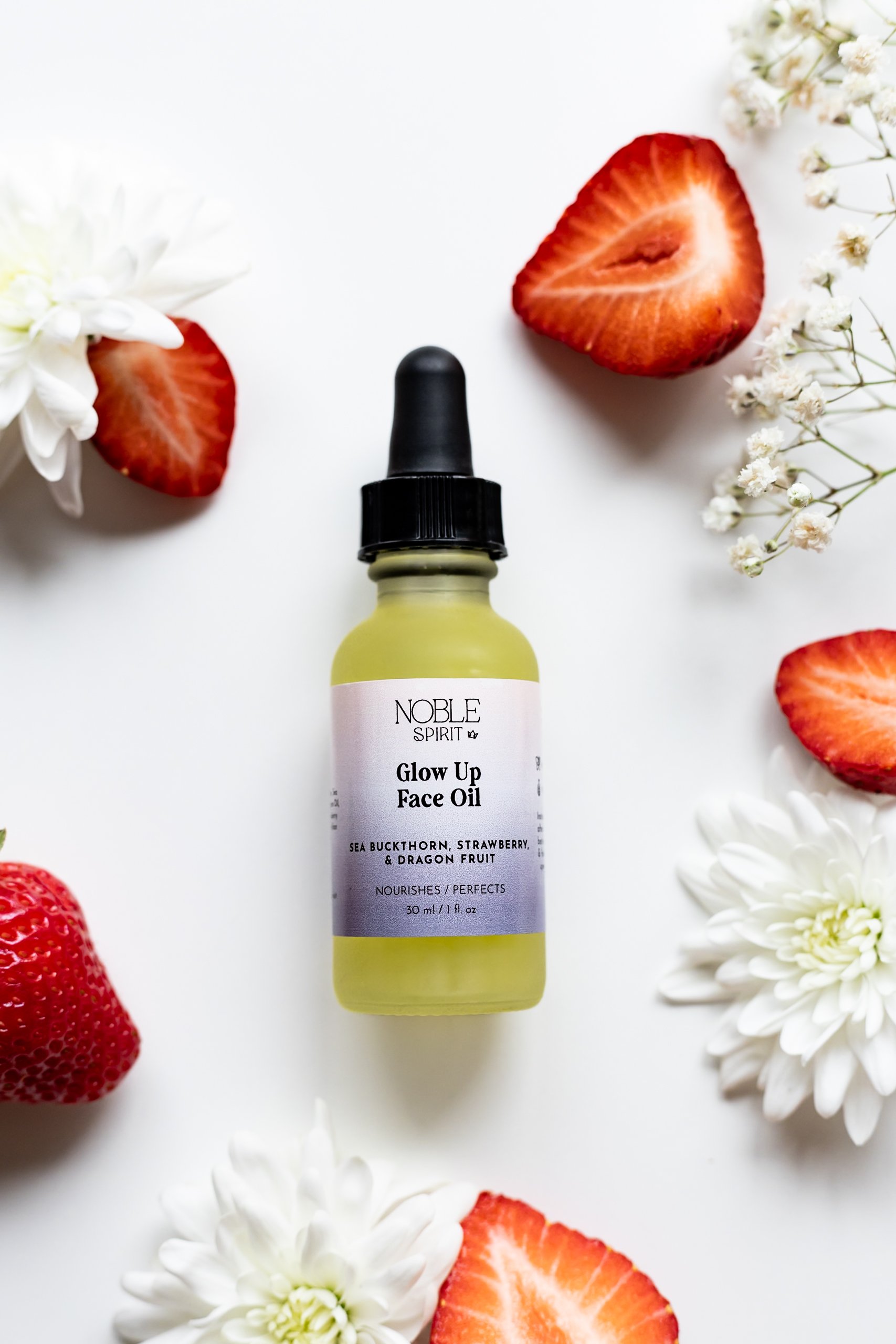 A bottle of Noble Spirit Glow Up Face Oil surrounded by sliced strawberries, white flowers, and baby's breath on a white background.