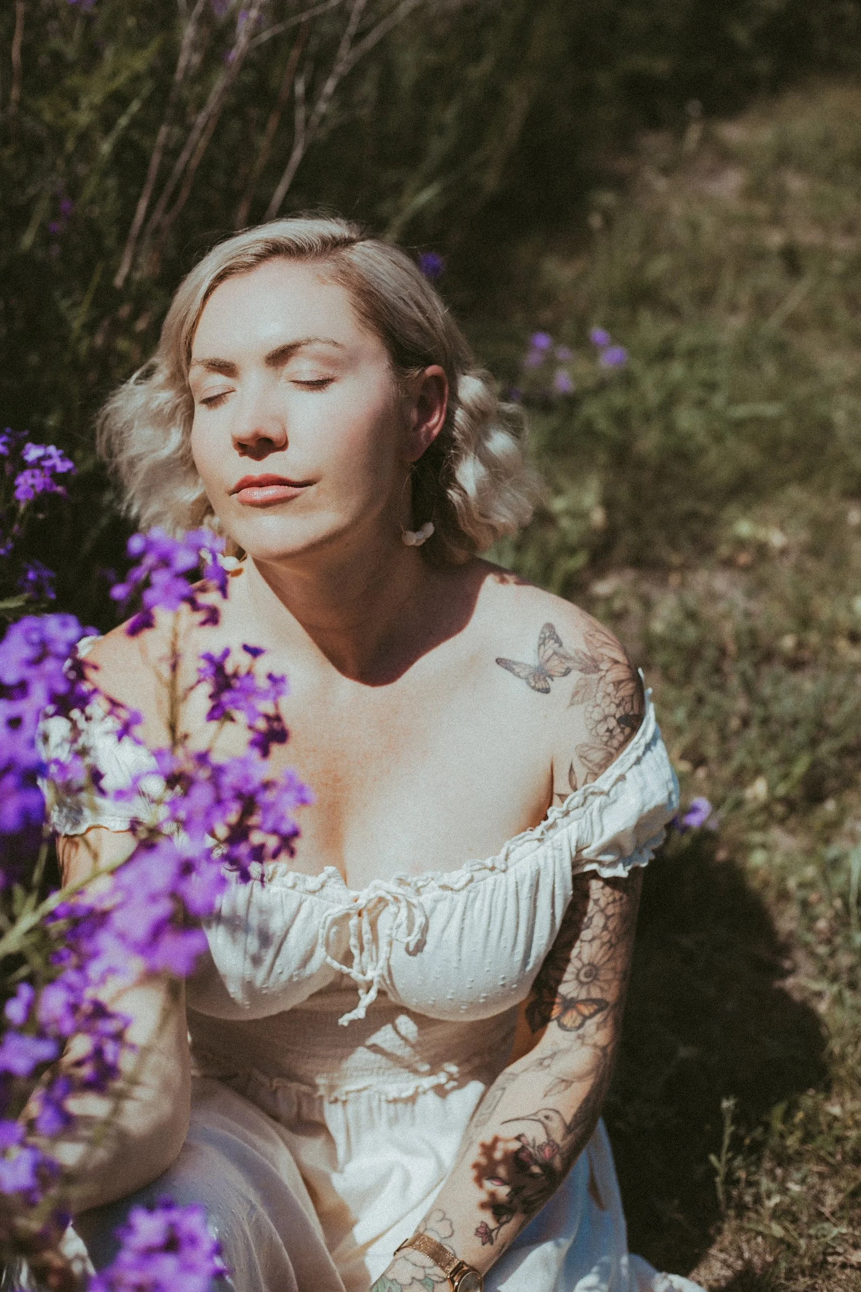 A woman with short blonde hair and tattoos on her arms is sitting outdoors with closed eyes, surrounded by purple flowers, wearing a white off-shoulder dress.