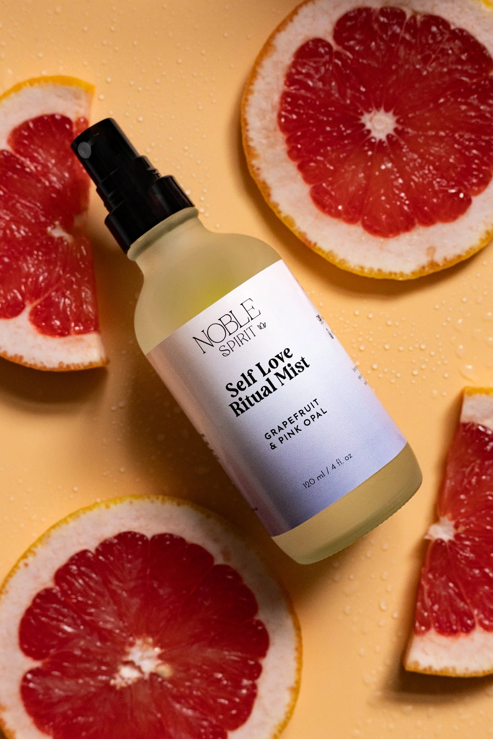 A bottle of Noble Spirit Self Love Ritual Mist surrounded by sliced grapefruits on a yellow background with water droplets.