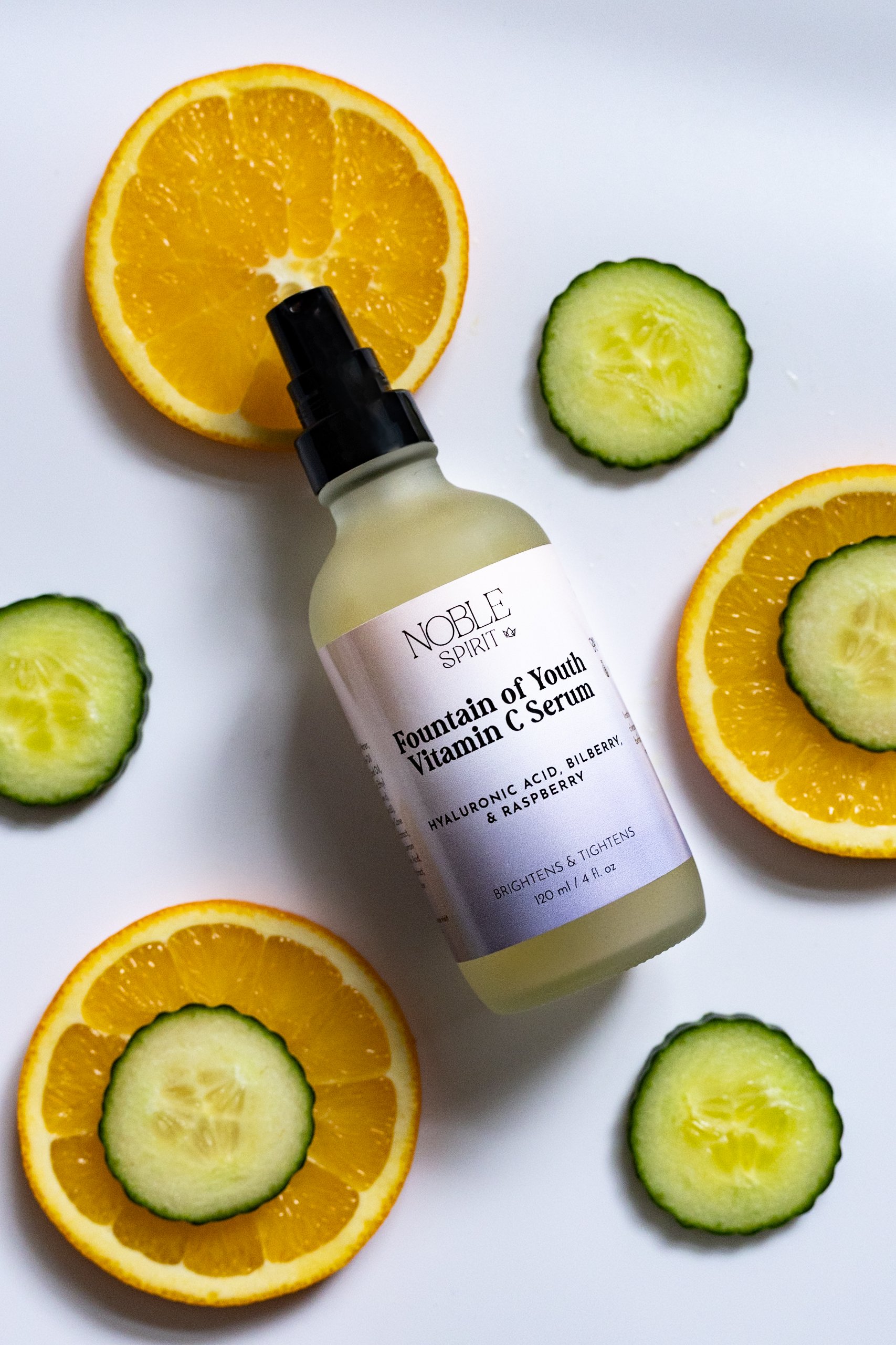 A bottle of Noble Spirit Fountain of Youth Vitamin C Serum surrounded by sliced cucumbers and oranges on a white surface.