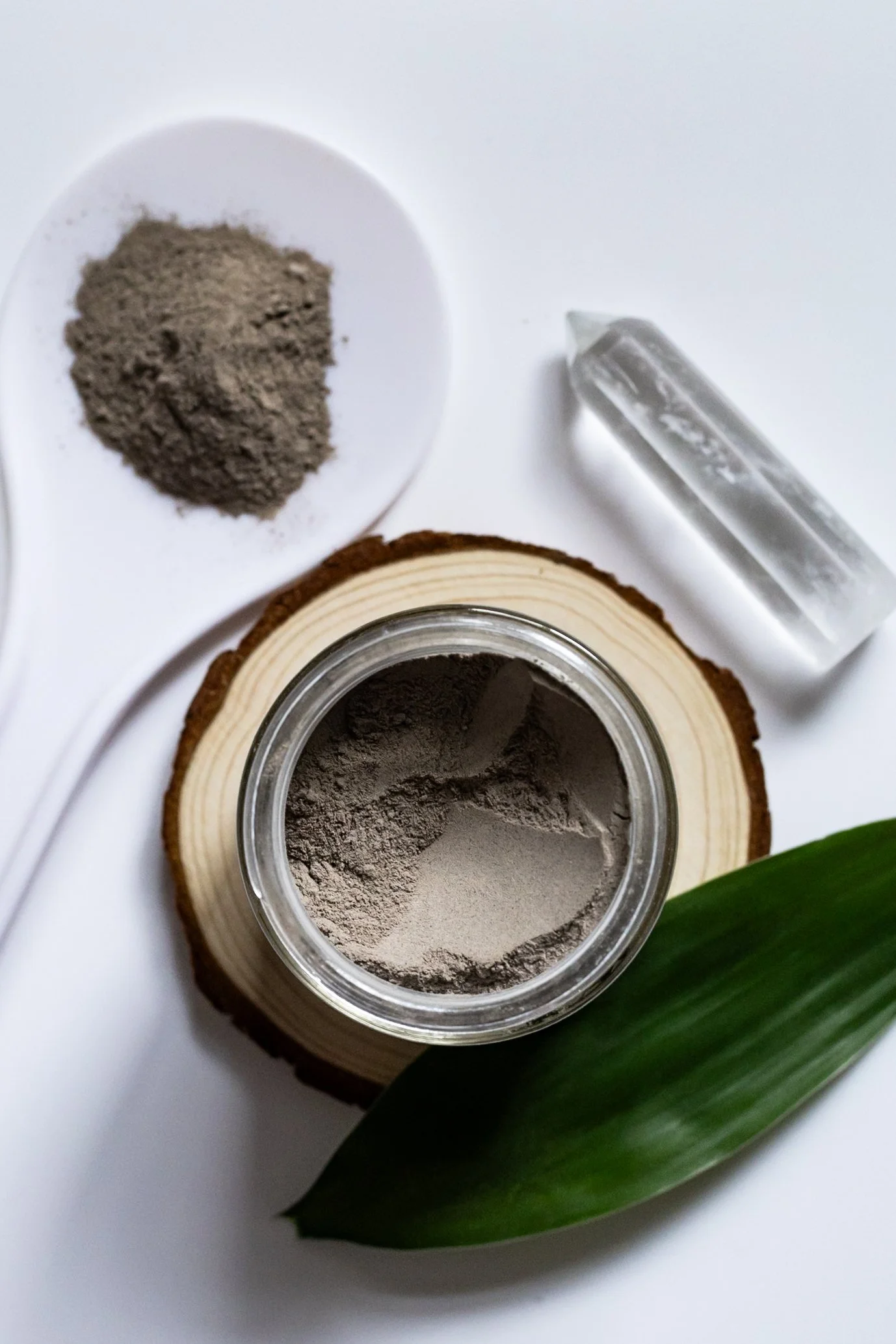 Open jar of gray powder, small pile of gray powder on a white dish, clear plastic container, green leaf on a wooden slab