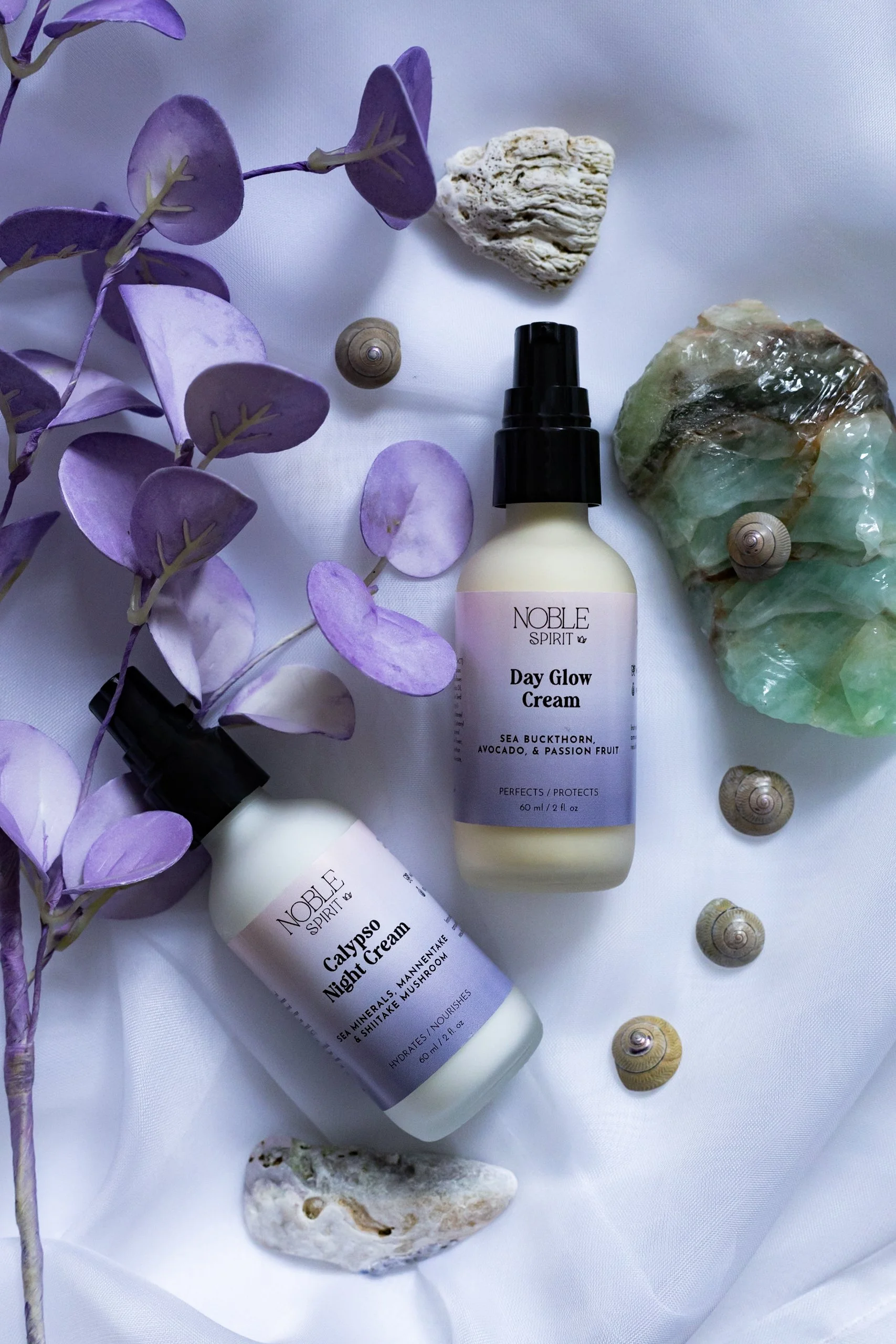 Skincare products with seashells and purple leaves on white fabric, including Noble Spirit Day Glow Cream and Calypso Night Cream.