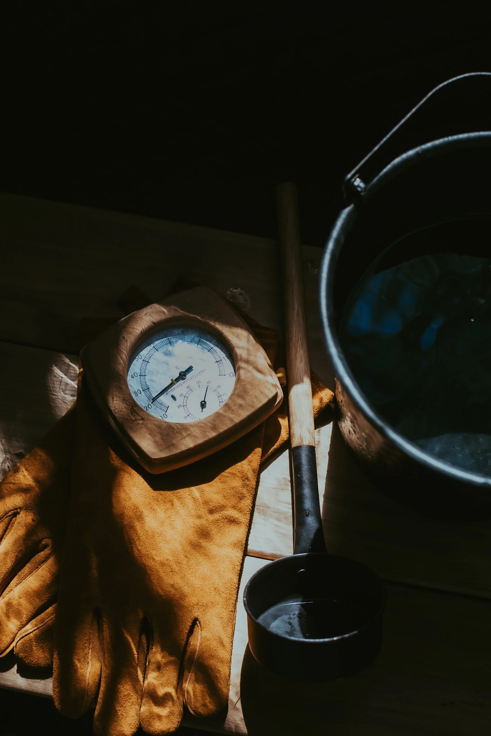 A thermometer, a pair of work gloves, a wooden handle tool, a metal bucket, and a small black container on a wooden surface, with sunlight casting shadows.