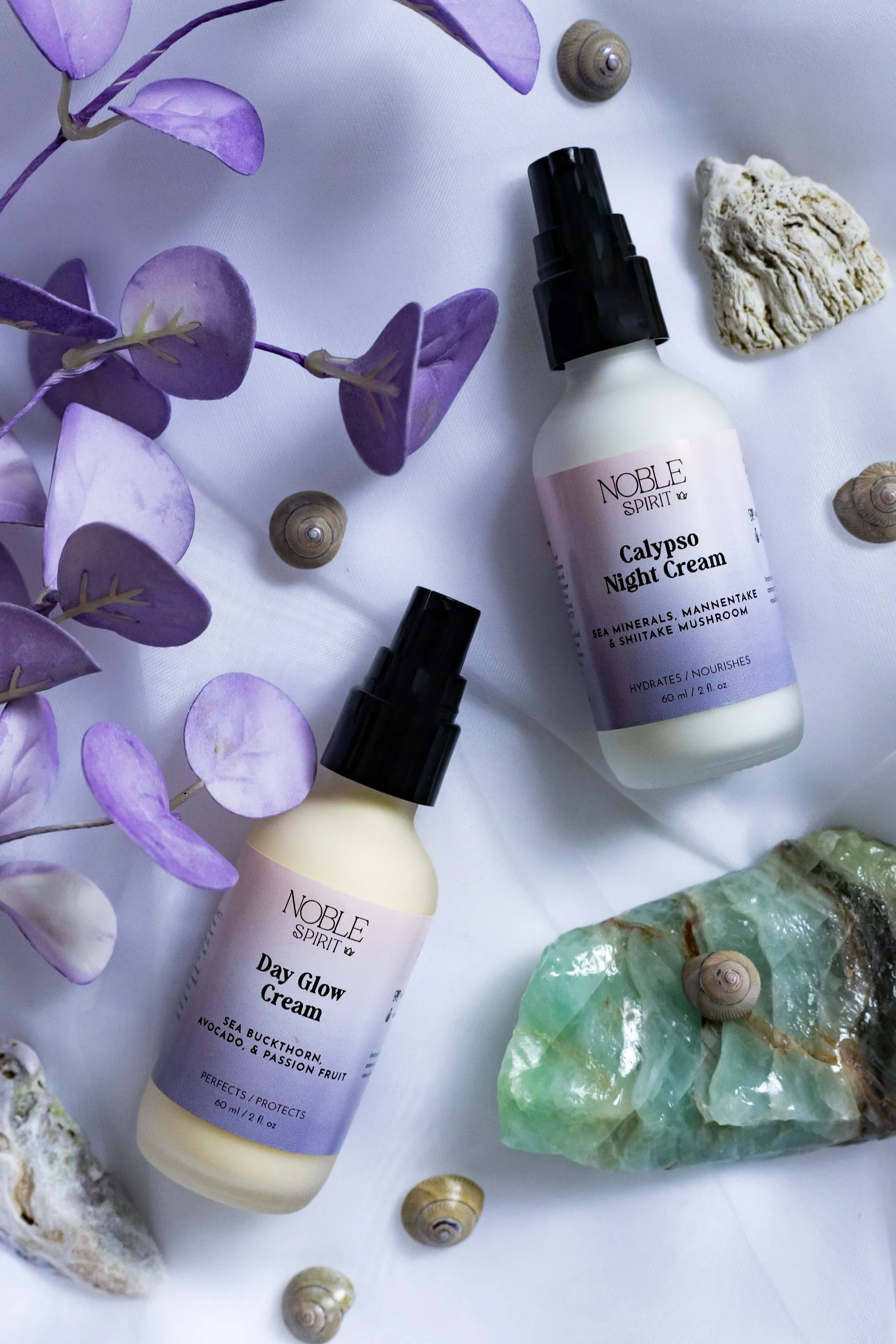 Two bottles of Noble Spirit skincare products, Calypso Night Cream and Day Glow Cream, surrounded by purple leaves, seashells, and stones on a white background.