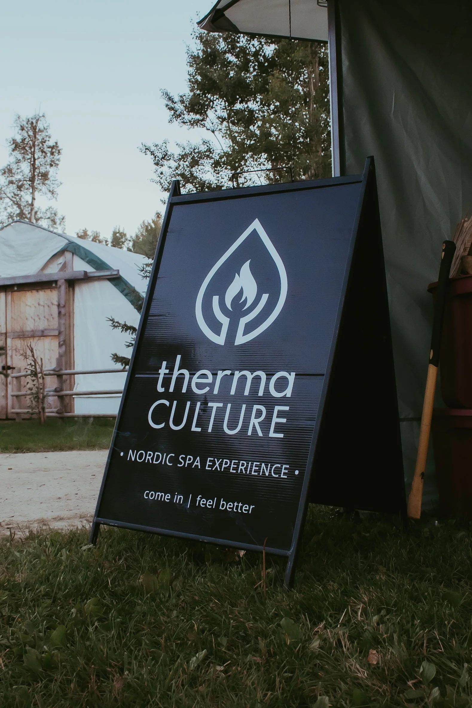 Outdoor sign for ThermaCulture Nordic Spa Experience with a logo of a stylized flame inside a water droplet. The sign reads: 'come in | feel better' and is placed on a grassy area near tents and trees.
