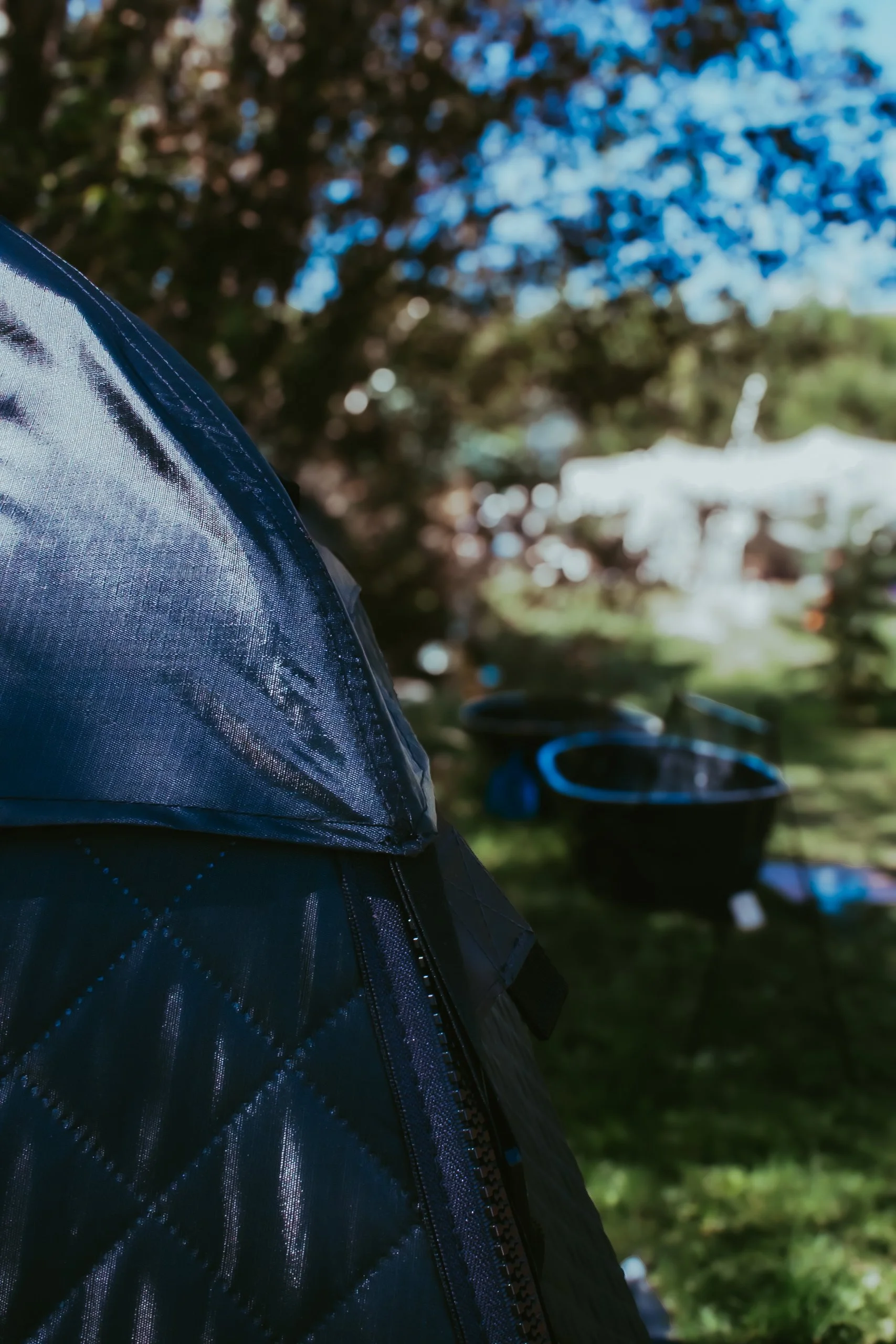Close-up of a dark blue quilted jacket with a shiny shoulder detail, with a blurred outdoor scene in the background showing green grass, tables, and umbrellas under a blue sky.