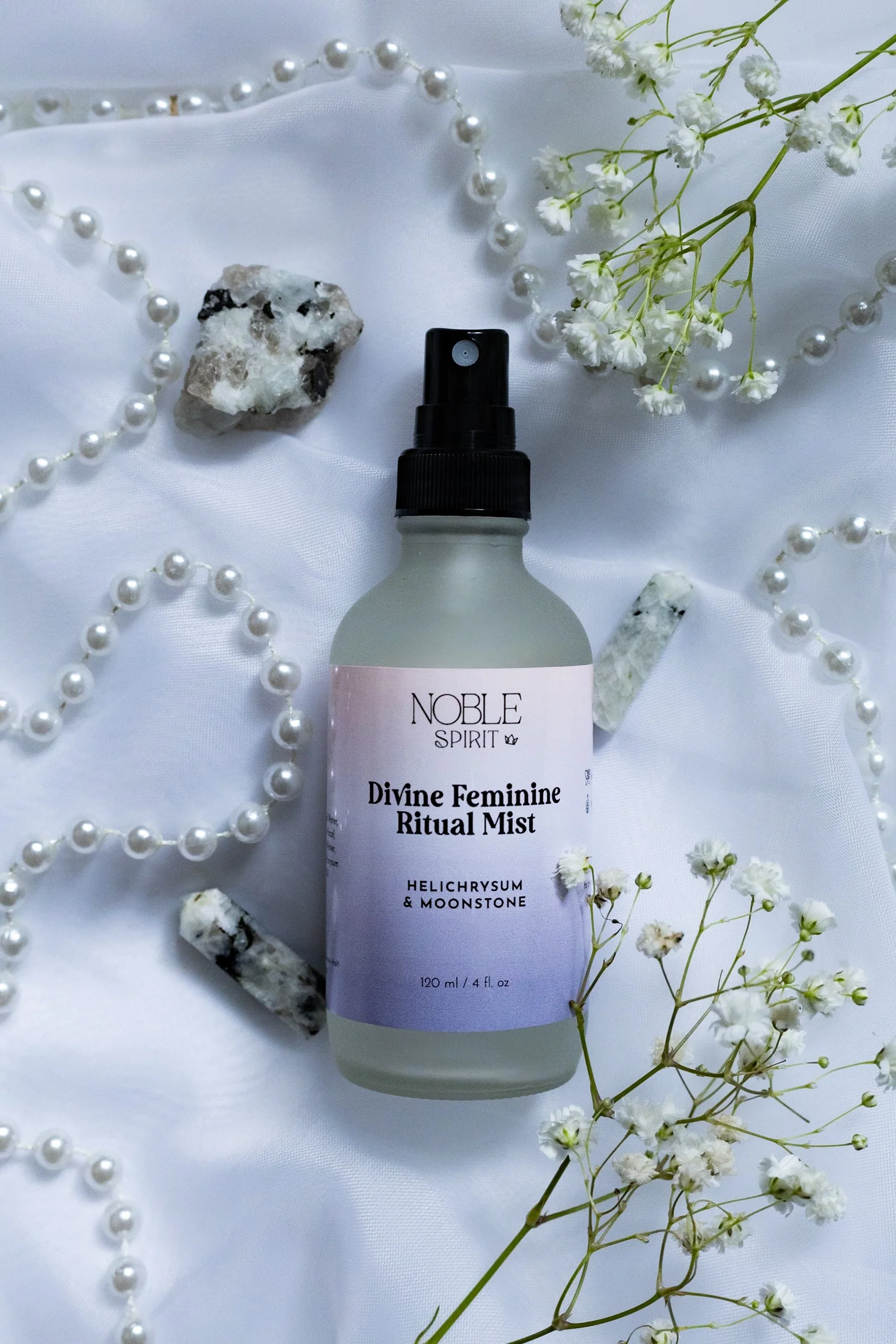 A spray bottle of Noble Spirit Divine Feminine Ritual Mist surrounded by white flowers, pearl necklace, and small rocks on white fabric.