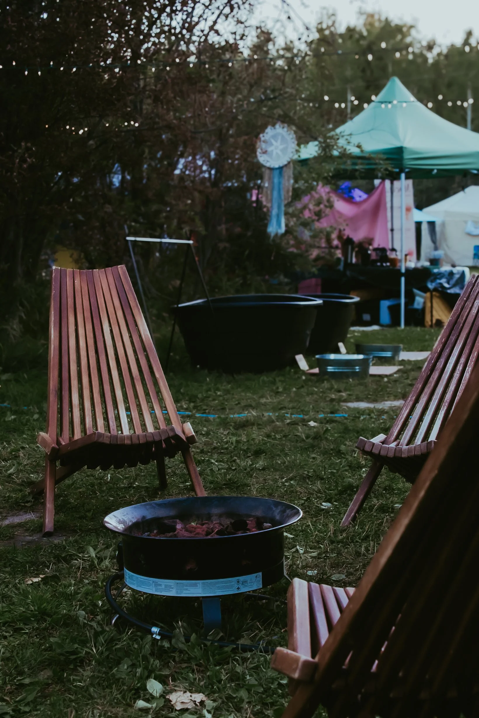 A backyard gathering setup with a small round fire pit, three wooden chairs, and a portable sauna tent in the background. String lights are hanging overhead, and there are large black containers and various items near the tent.