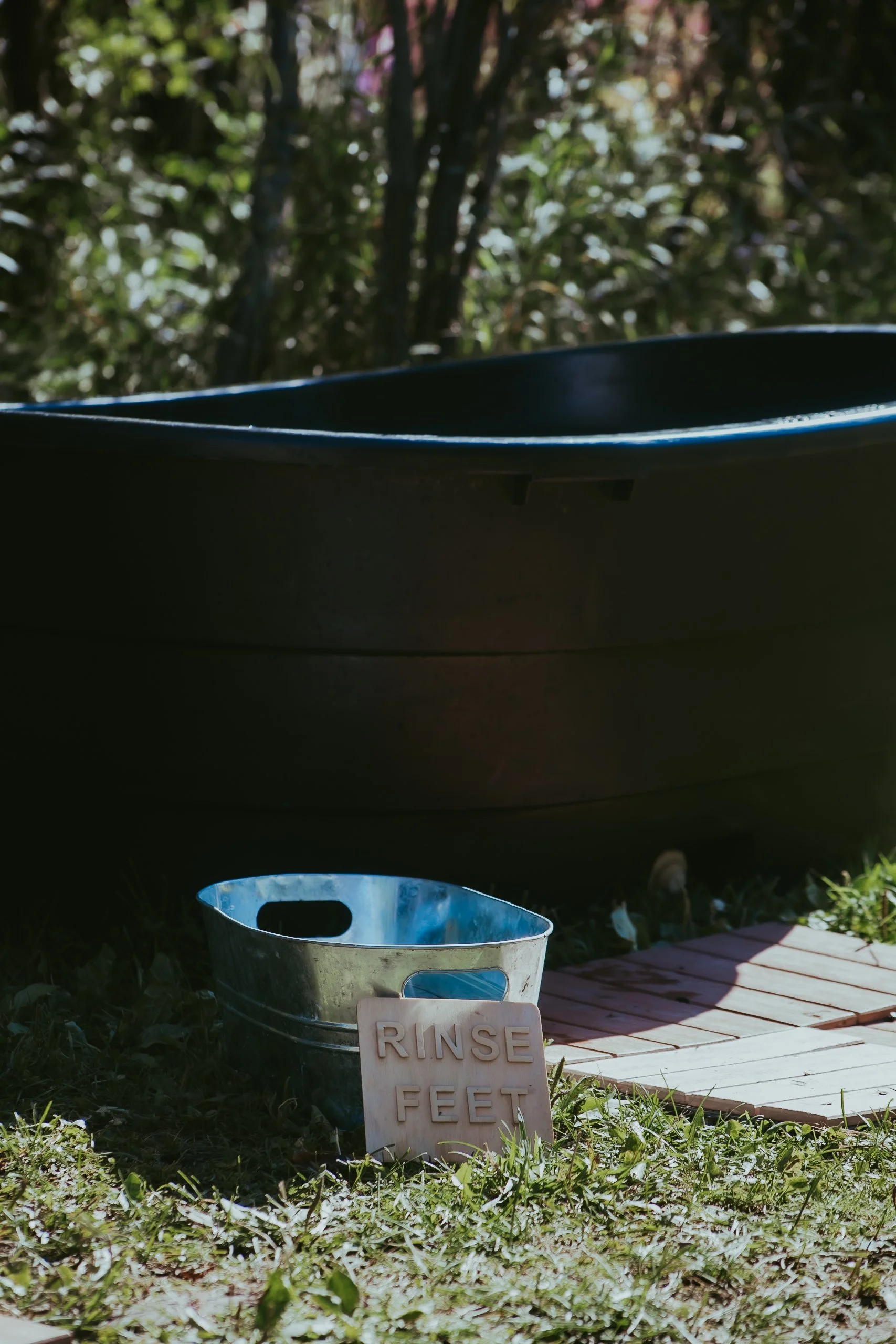 A black outdoor tub with a sign that says "RINSE FEET," a metal bucket, and a wooden mat on grass, with trees in the background.