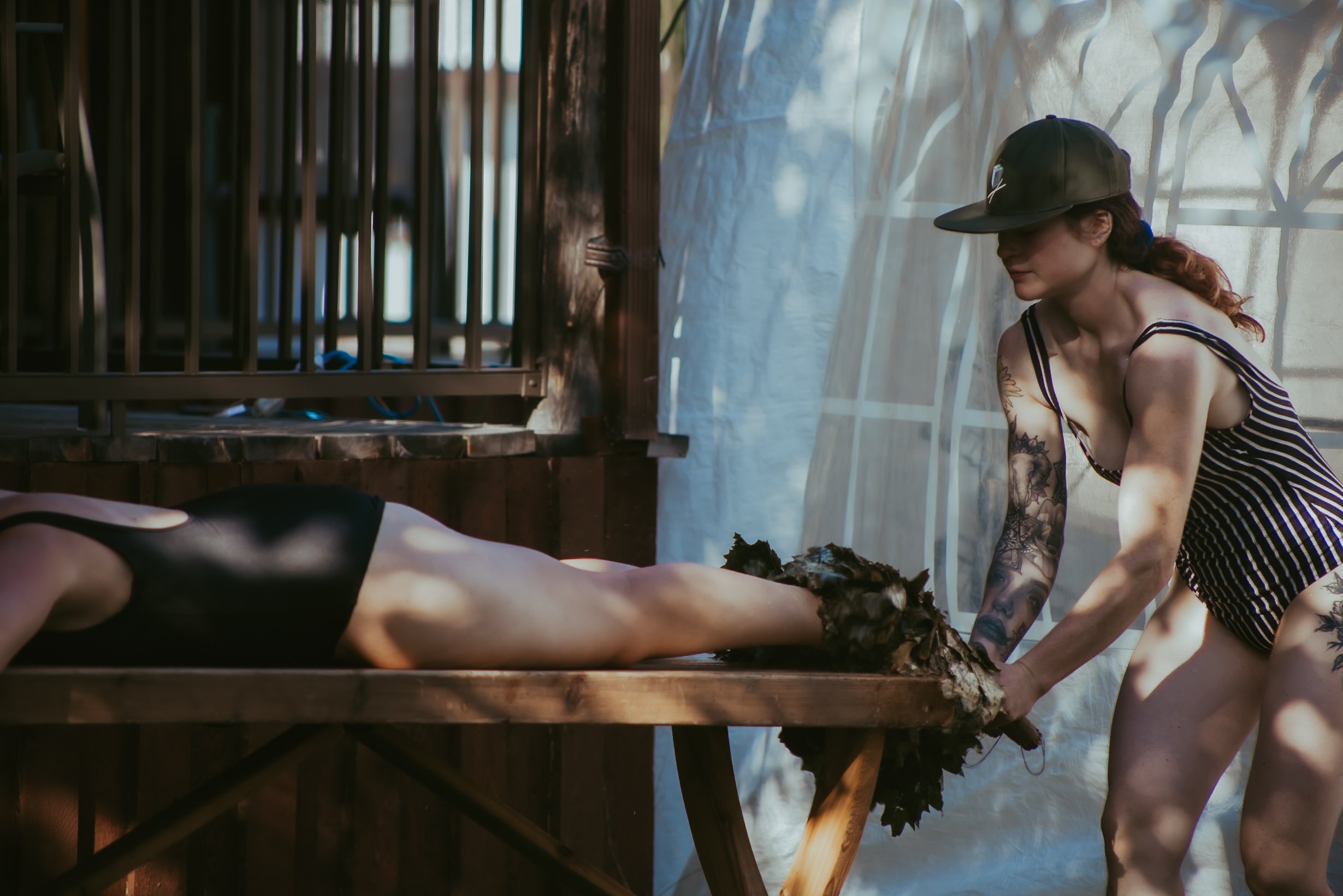 A woman with tattoos covering a persons feet with banana leaves to clear out old energy while the person is lying face down on massage table, located outdoors.