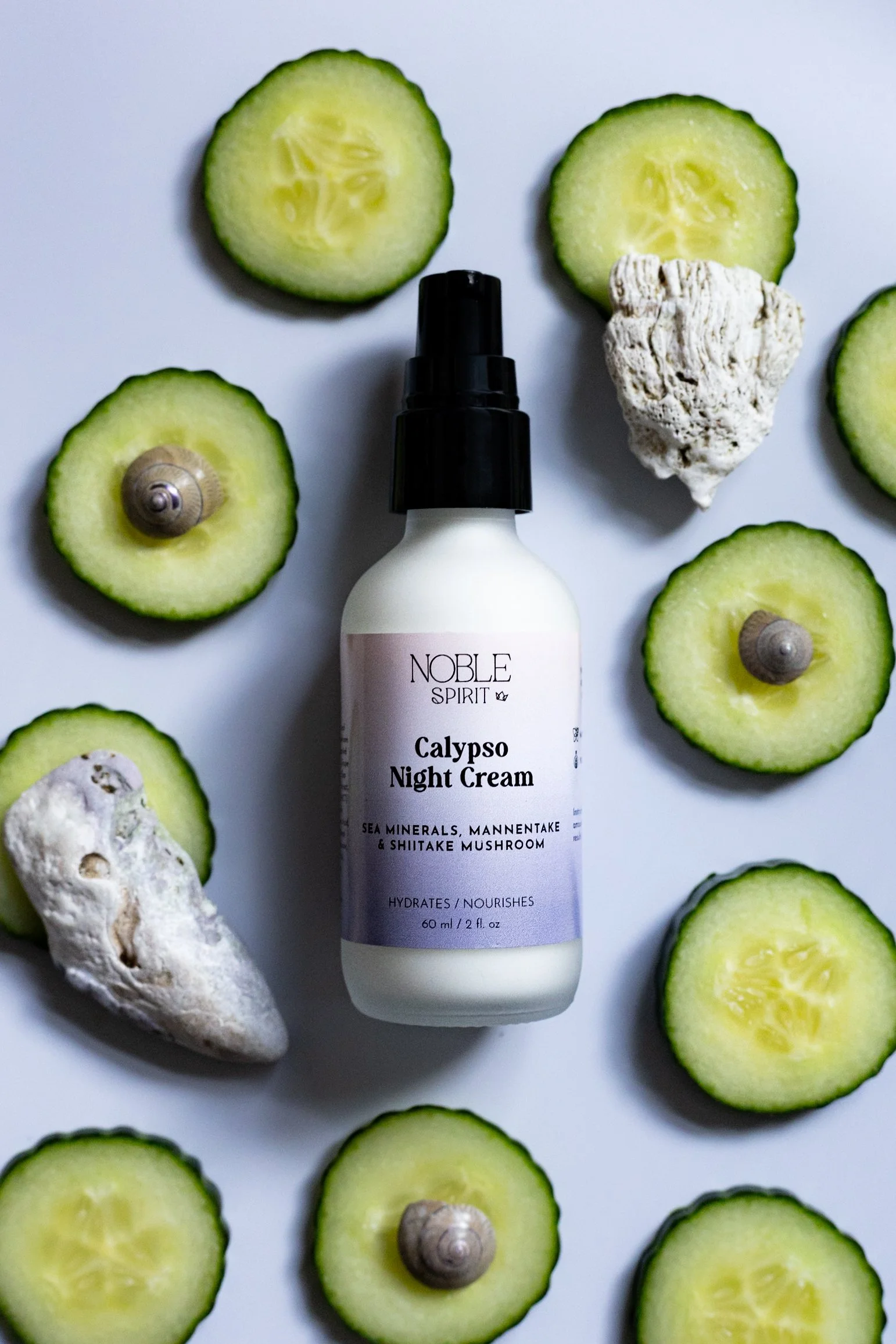 Bottle of Noble Spirit Calypso Night Cream surrounded by cucumber slices, seashells, and rocks on a white surface.