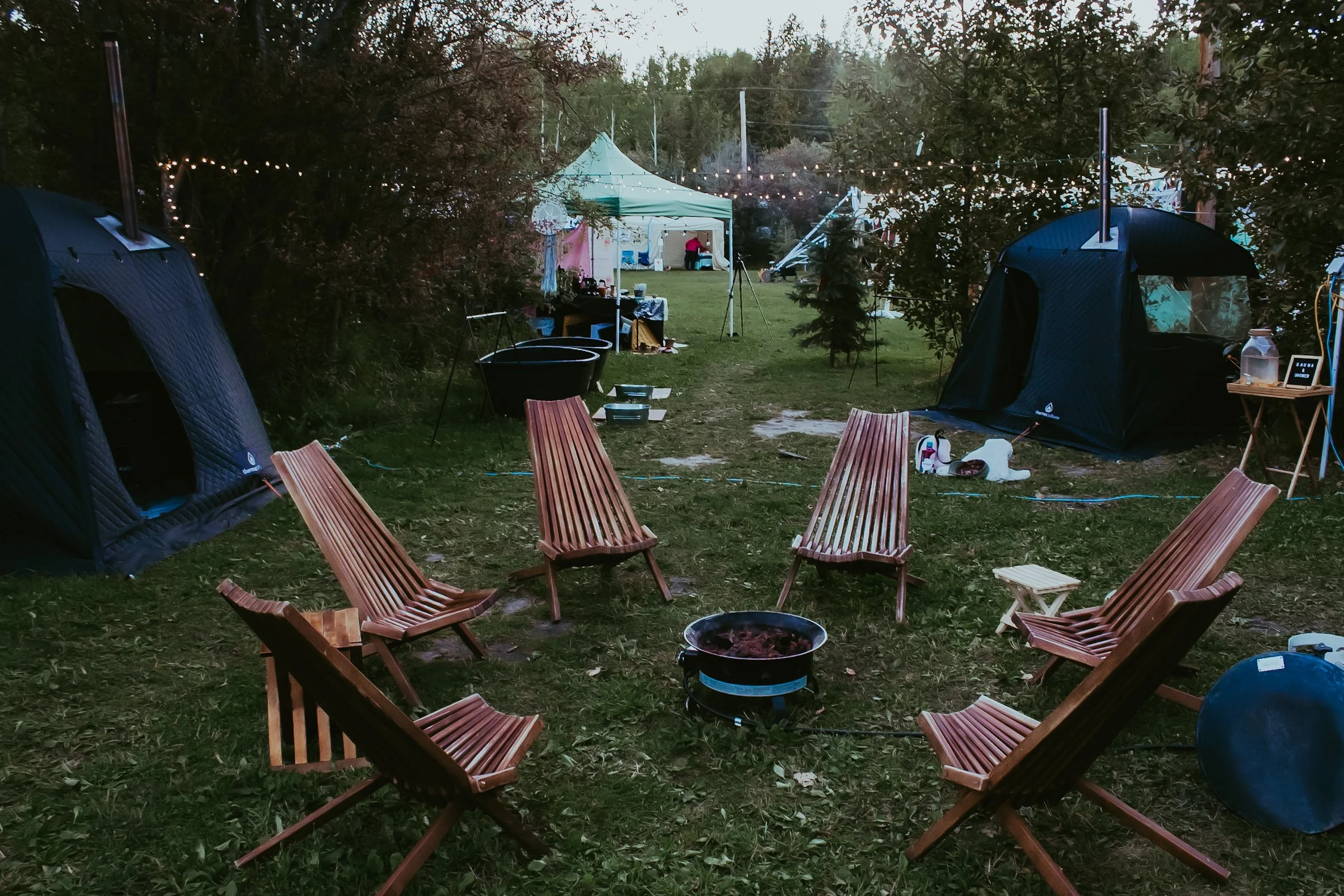 Outdoor camping scene with six wooden chairs arranged in a circle around a fire pit, tents, string lights, and various camping supplies in a wooded area.