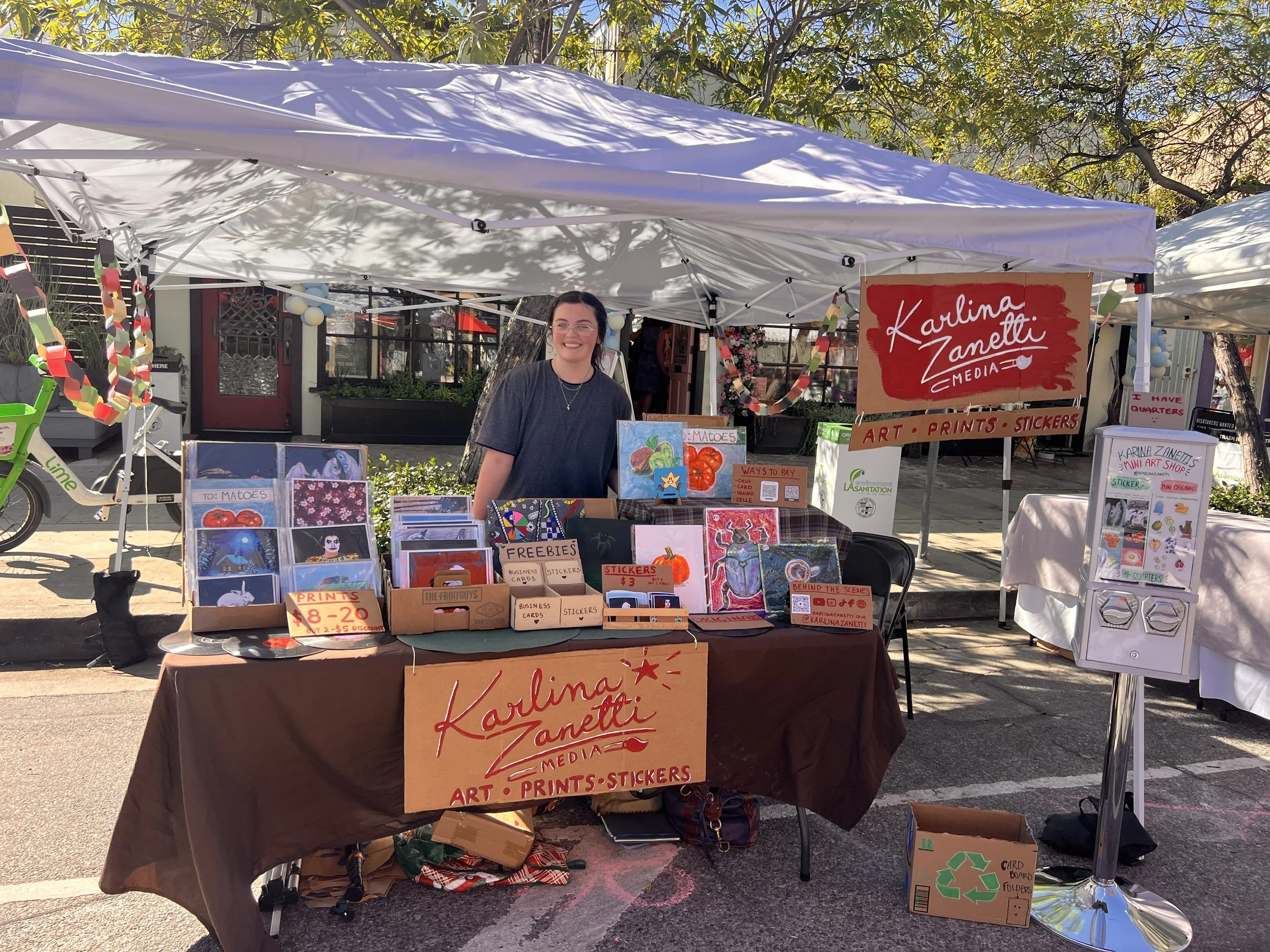 A woman with glasses and a dark T-shirt standing at a booth selling art, prints, stickers, and mini art. The booth has a large sign reading 'Karina Zanetti Media.' There are various artwork and cards displayed on the table. The booth is set up outdoors under a white canopy, with trees in the background.