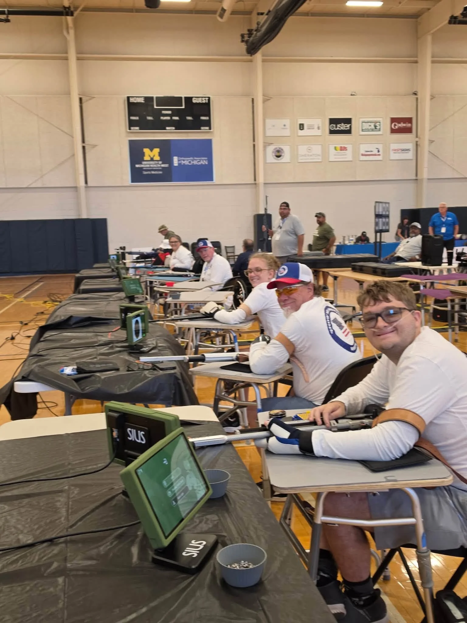 People sitting at tables in a gymnasium, participating in an event with laptops and electronic devices. The background shows a scoreboard and banners with logos.