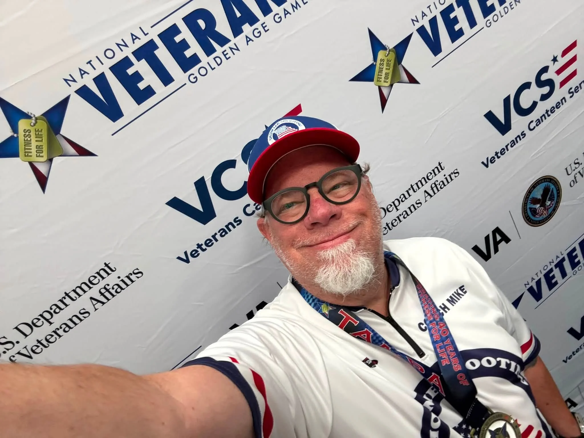 Smiling man wearing glasses, a white shirt with patriotic designs, a red cap, and a lanyard with a medal, taking a selfie in front of a Veterans Affairs banner.