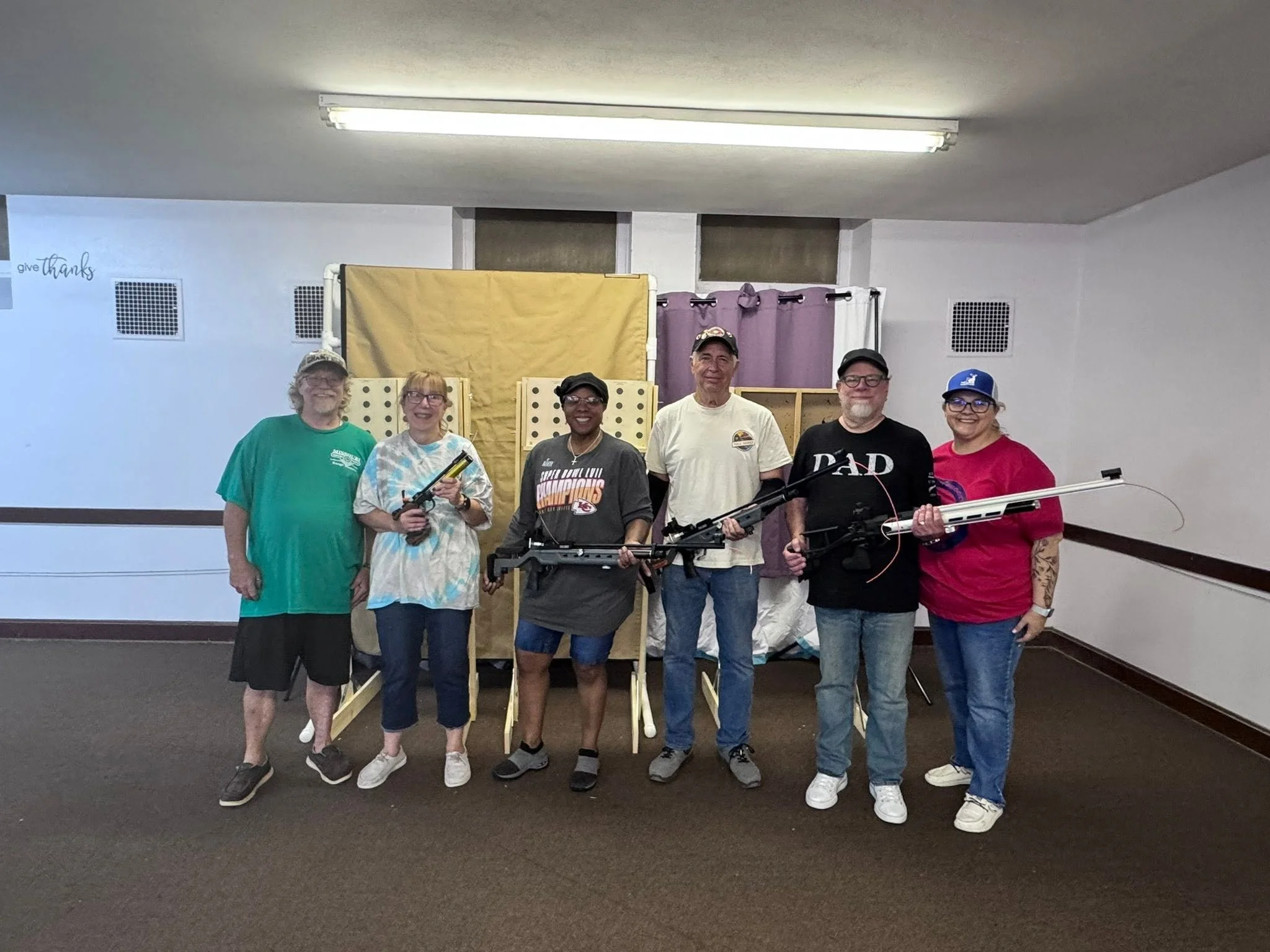 Group of seven people standing indoors, smiling, with a yellow and purple barrier behind them, and holding Bows, in a room with white walls and fluorescent ceiling lights.