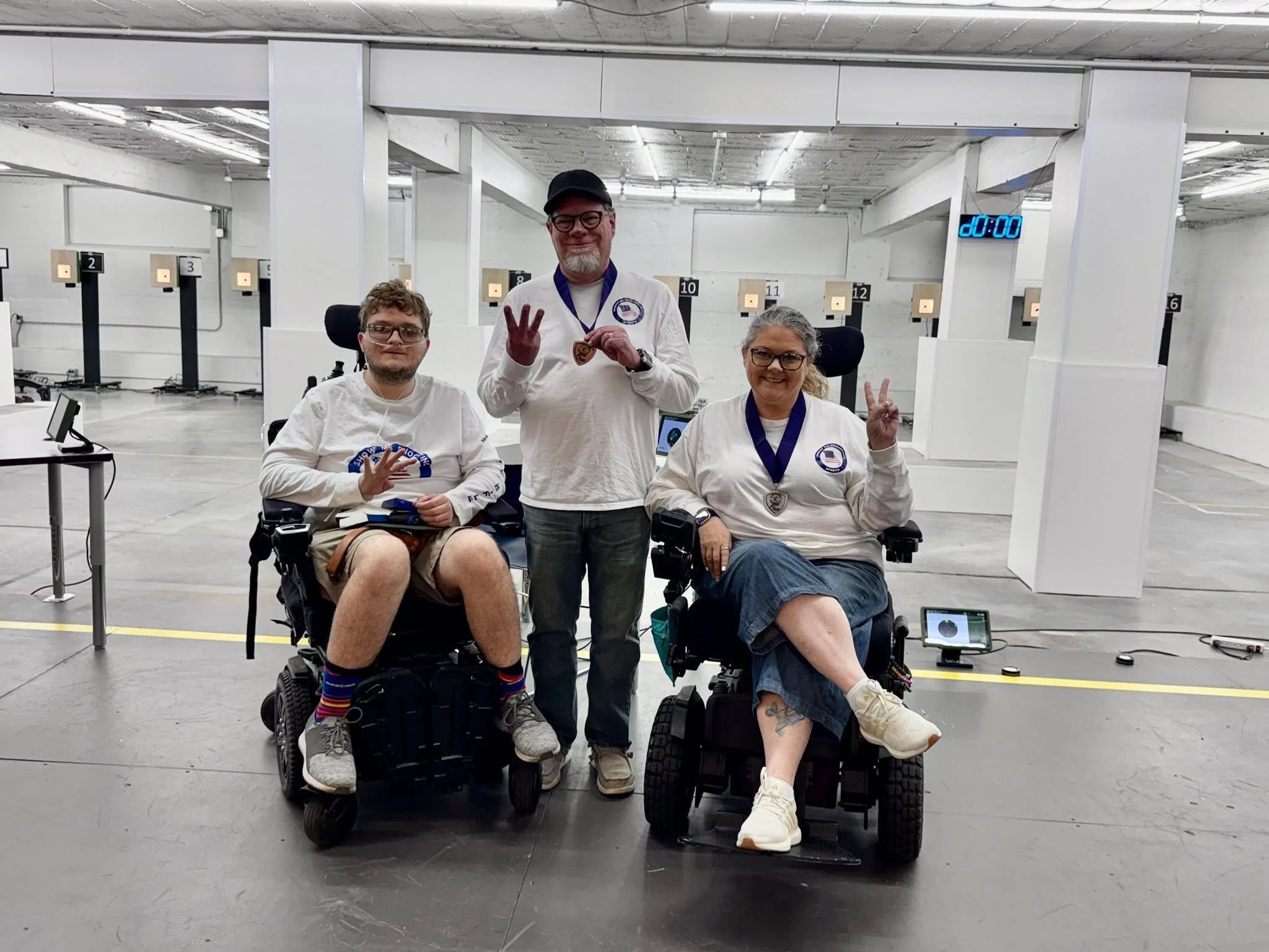 Group of three individuals, two in wheelchairs and one standing, in a large indoor shooting range with target stations in the background. The three are smiling and showing peace signs, with medals around their necks.
