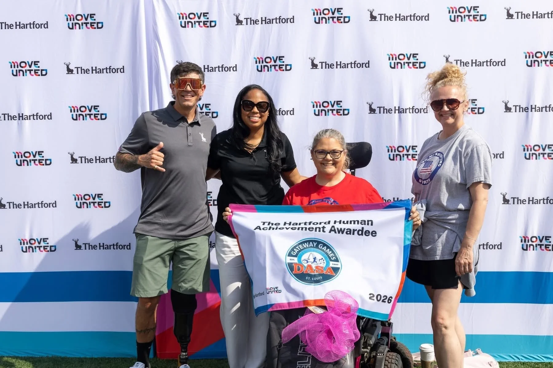 Group of four diverse people standing in front of a backdrop with the words "MOVE UNITED" and "The Hartford," holding a banner that reads "The Hartford Human Achievement Award" and a logo for Gateway Games DASA, with a young woman in a wheelchair in the center, smiling.