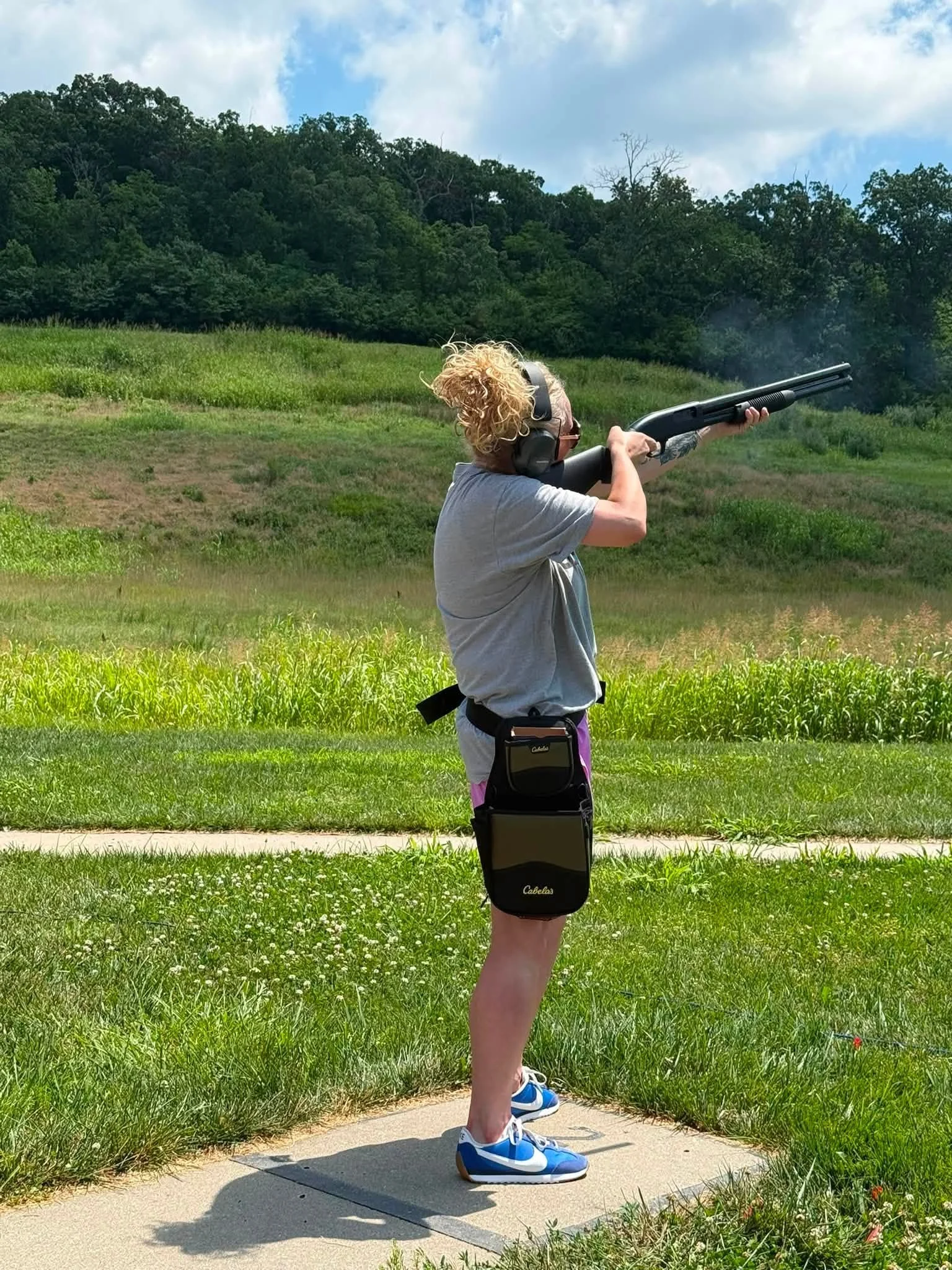 Person with curly blonde hair wearing headphones, a gray t-shirt, and sneakers, aiming a shotgun outdoors on a concrete pad, with green grass, foliage, and trees in the background under a partly cloudy sky.