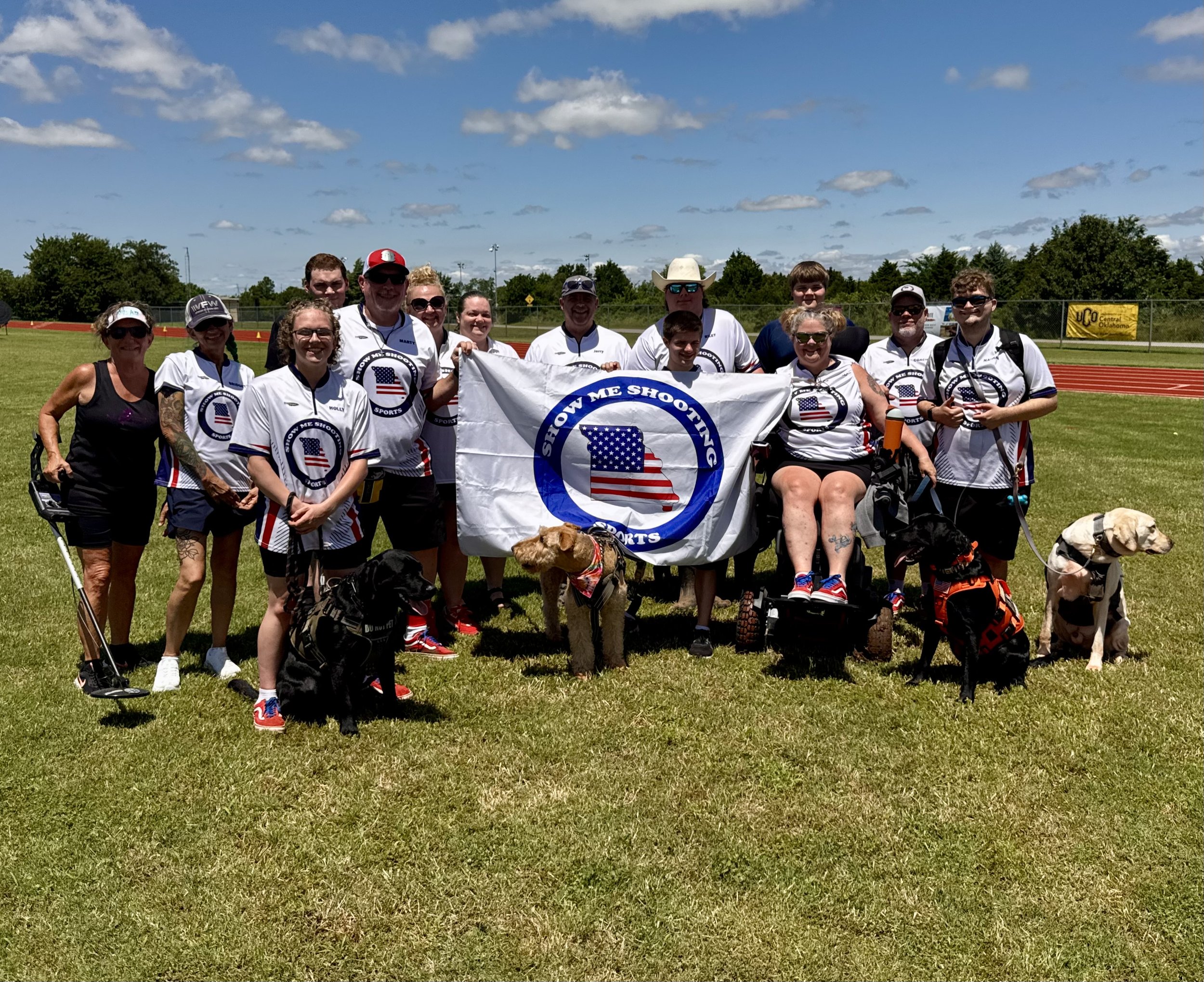 A group of people, some wearing sports jerseys, holding a banner that says 'Show Me Shooting' with a logo of the state of Missouri and American flag. They are outdoors on a sports field with cloudy blue sky and trees in the background.
