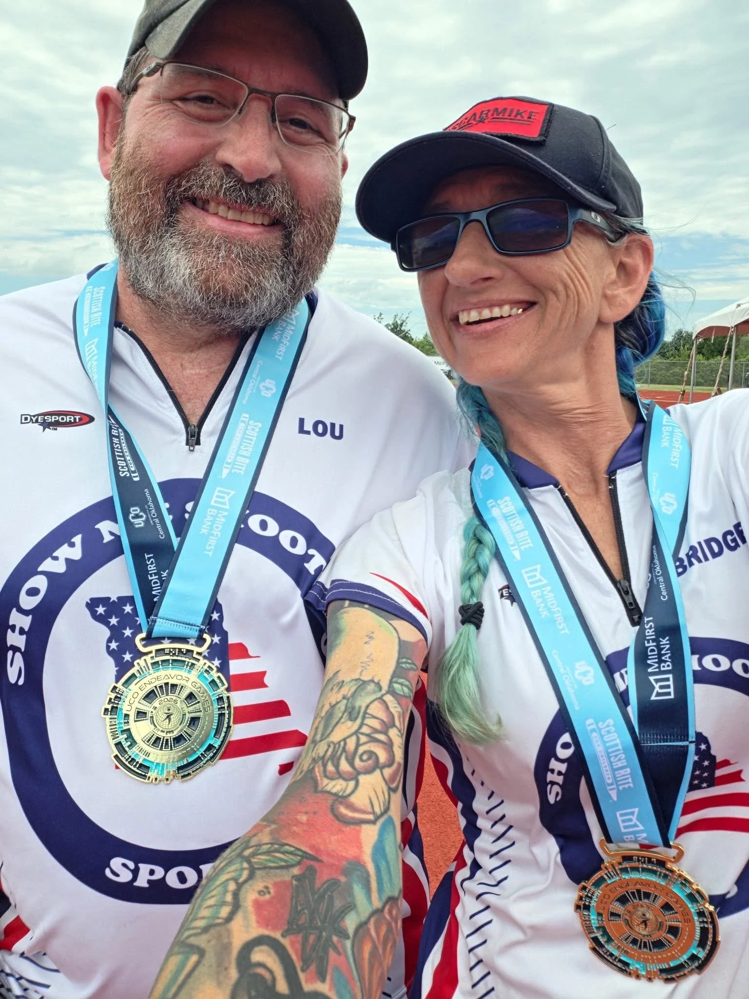 Two smiling people wearing medals and race shirts, taking a selfie outdoors on a cloudy day. Both wearing Show Me Shooting Jerseys.' They are wearing sunglasses and caps, and one has a colorful tattoo on his arm.