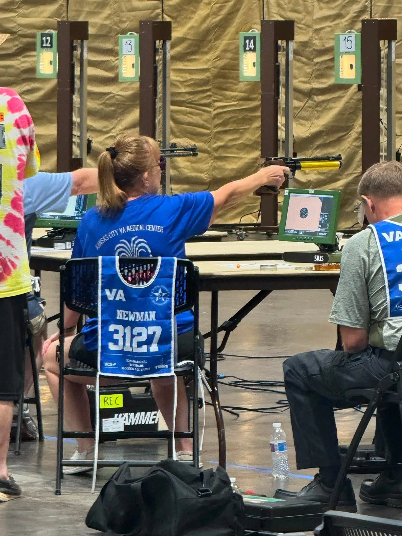 Person in wheelchair at shooting range aiming a handgun at targets, with people and equipment nearby.