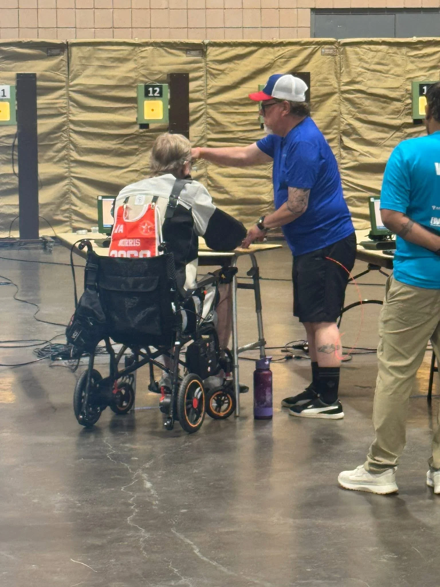 A man with tattoos, wearing a blue shirt, black shorts, and a red, white, and blue cap, adjusts the focus of an eye examination for a woman in a wheelchair. The woman has gray hair, glasses, and a beige and black jacket. They are in a room with a brick wall and yellow barriers behind them, with various electronic equipment on tables nearby.