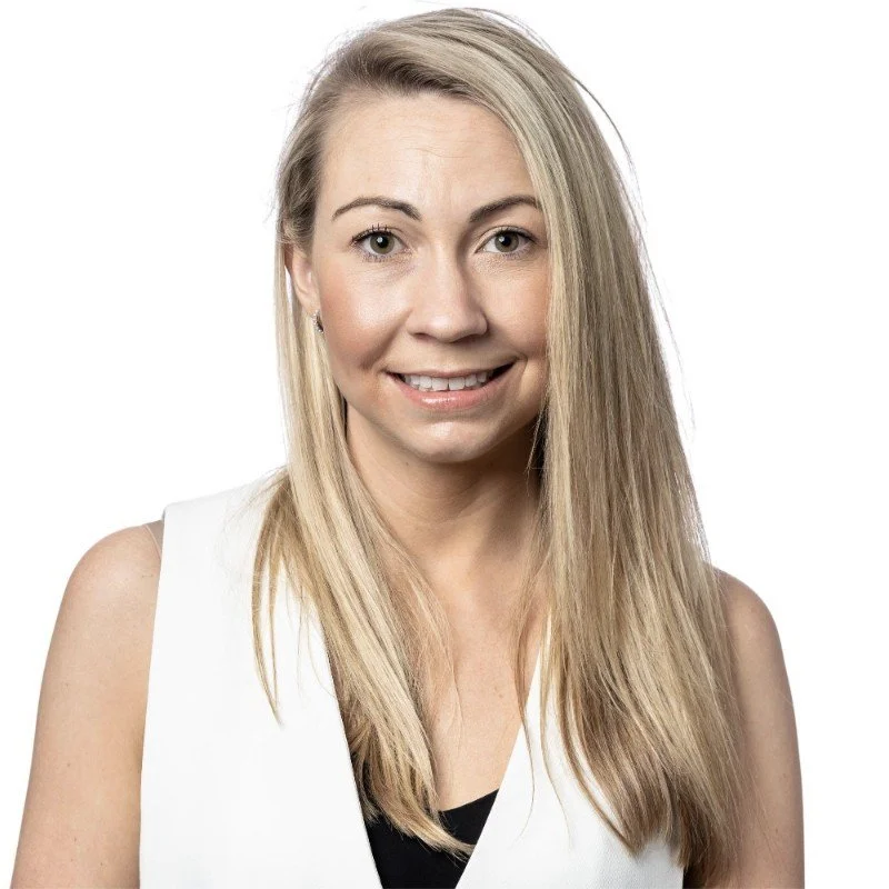 A woman with long blonde hair and light skin smiling at the camera, wearing a sleeveless white top against a white background.