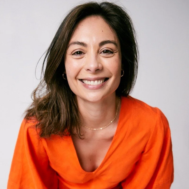 A woman with shoulder-length dark hair smiling, wearing an orange top and small gold hoop earrings, against a plain light grey background.